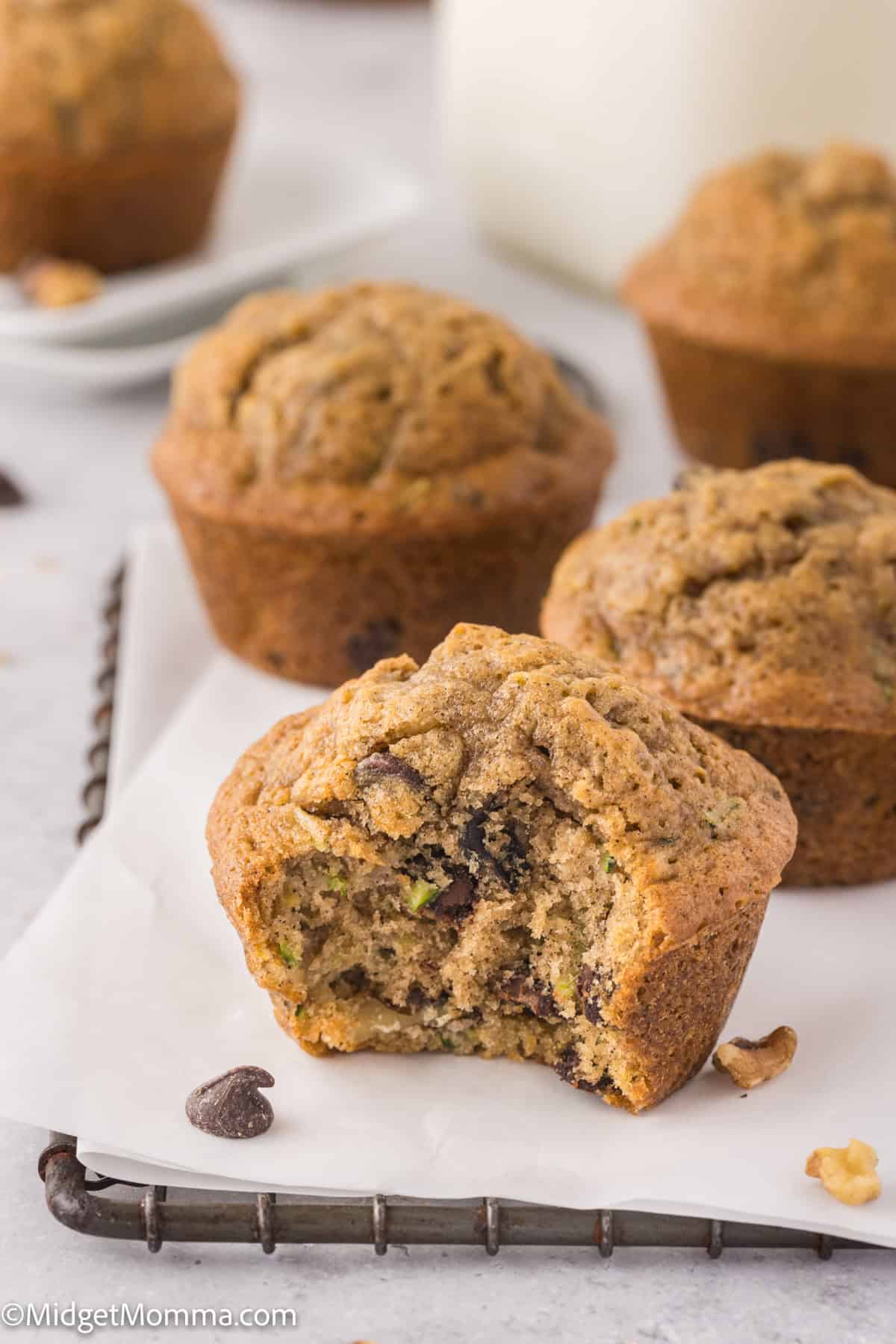 Close-up of chocolate chip muffins on parchment paper, with one muffin missing a bite. Pieces of chocolate and nuts are visible. Background shows a glass of milk.