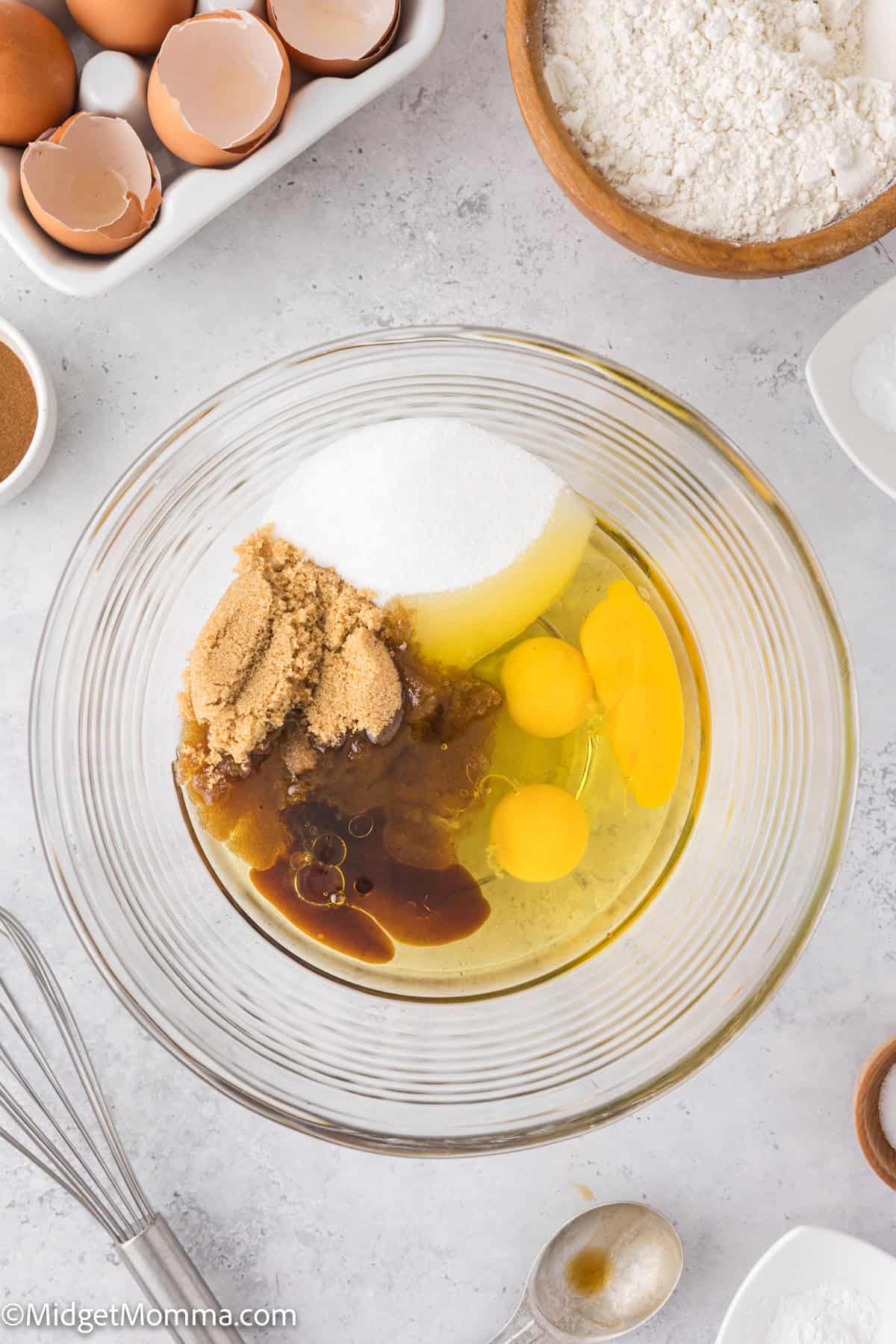 A mixing bowl with eggs, brown sugar, white sugar, and vanilla extract. Surrounding are flour, eggs, cinnamon, and a whisk.