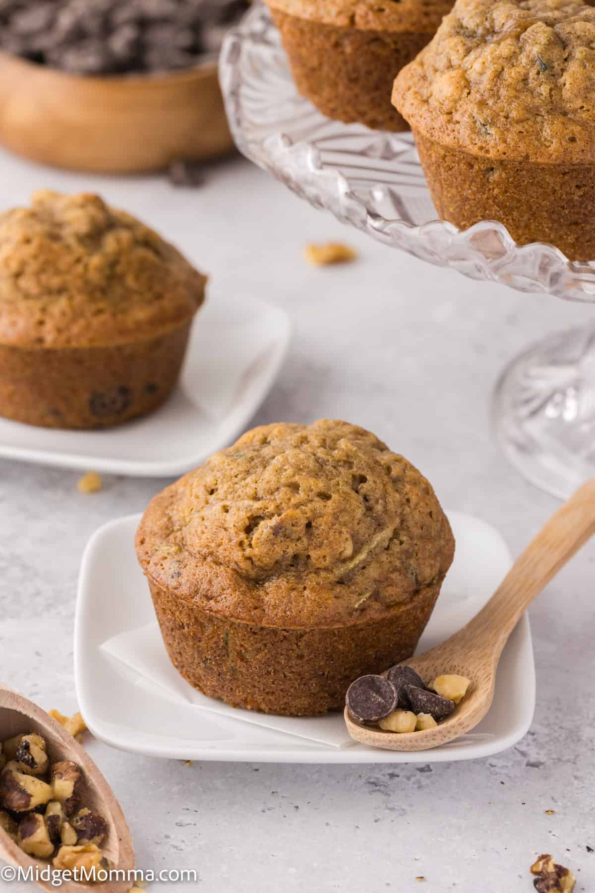 Banana muffins on white plates, with a wooden spoon holding chocolate chips and nuts. A glass stand holds additional muffins in the background.