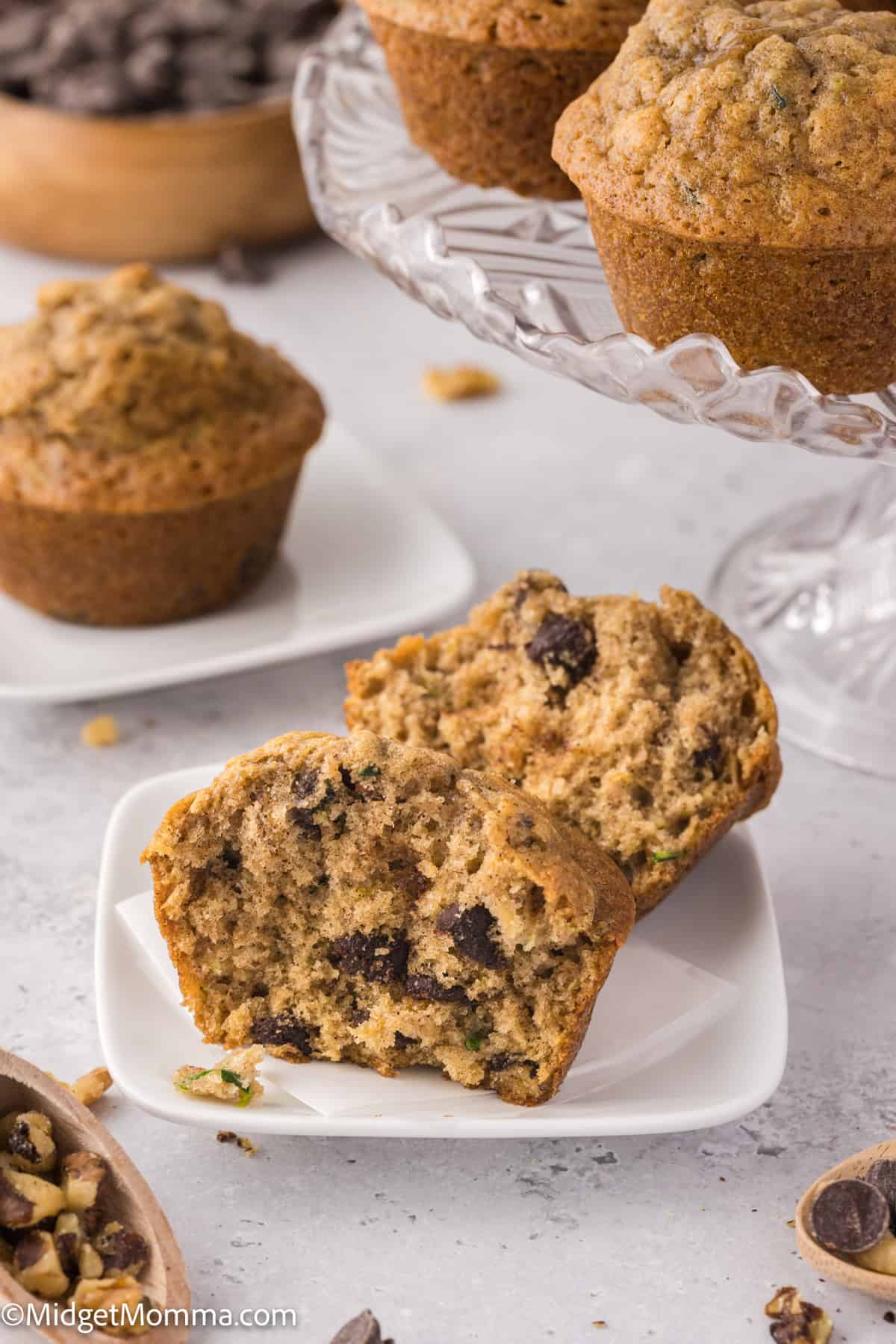 Close-up of a sliced chocolate chip muffin on a white plate, surrounded by whole muffins and scattered nuts.