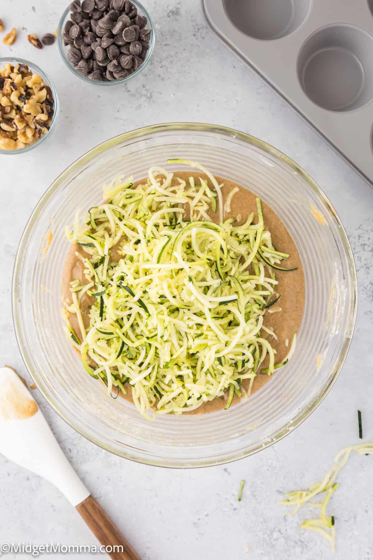 A glass bowl with grated zucchini mixed into a batter, a wooden spoon on the side, and nearby bowls containing chocolate chips and walnuts. A muffin tray is in the upper right corner.