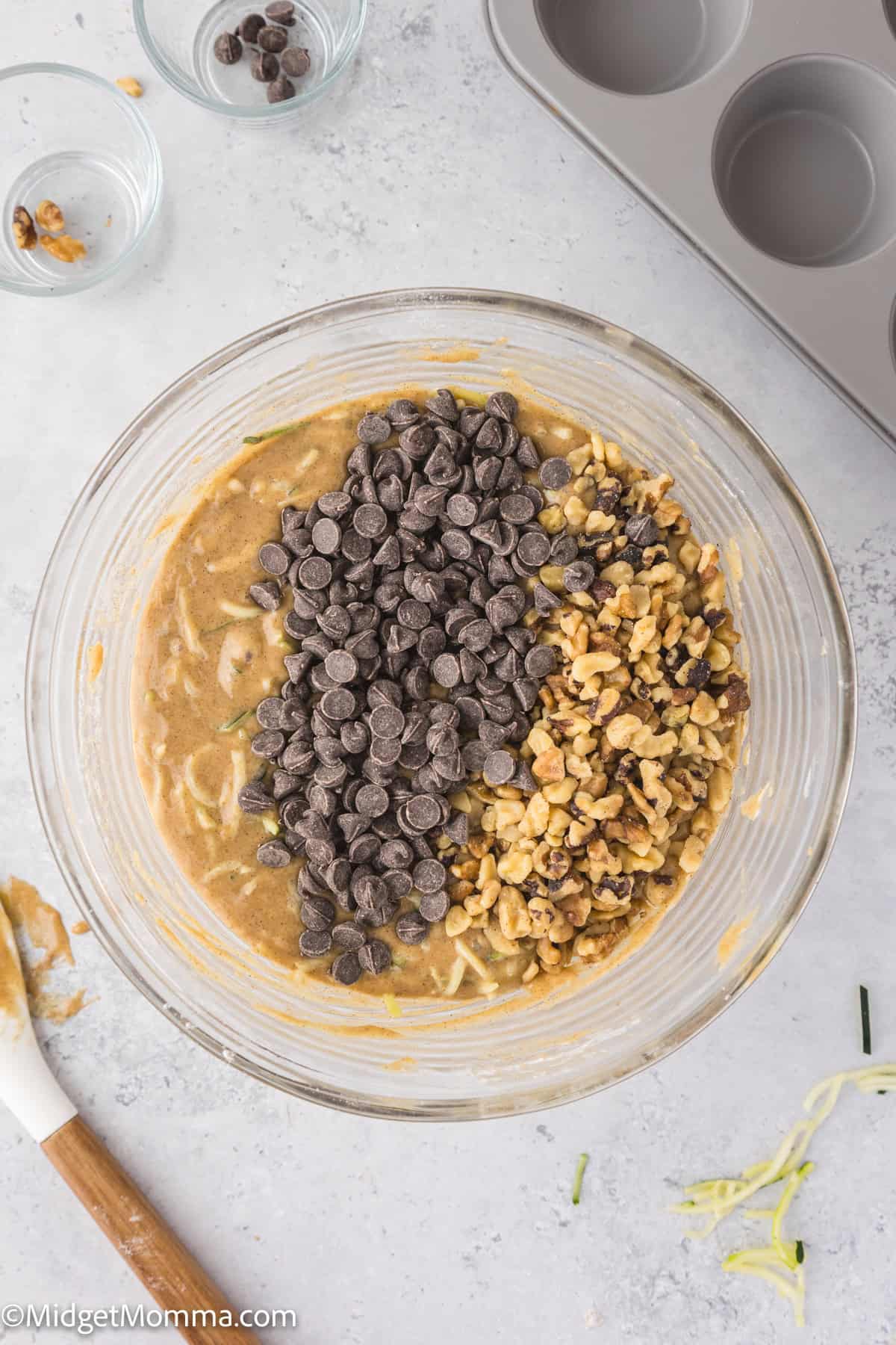 A mixing bowl with batter, chocolate chips, and chopped nuts. A rubber spatula is nearby, with empty bowls and a muffin tin in the background.