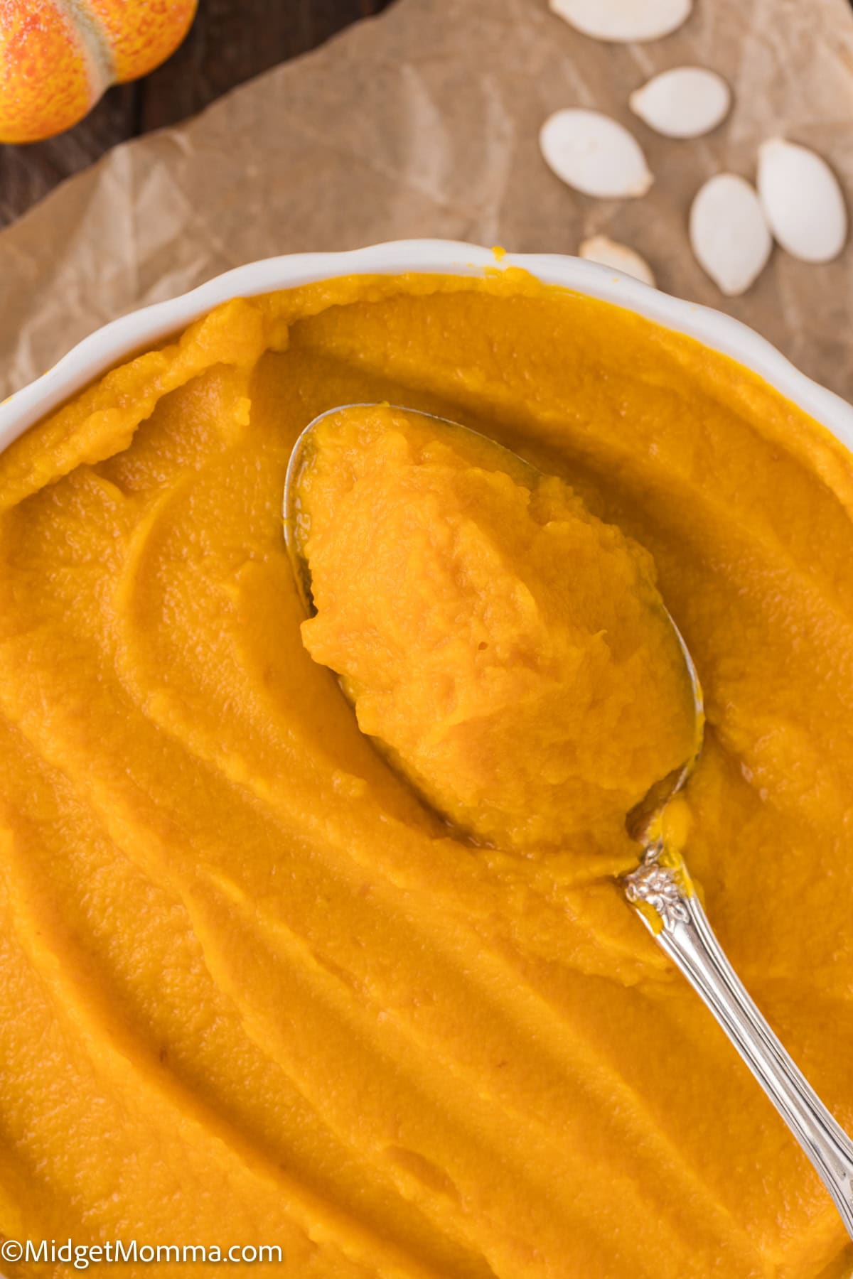 A bowl of smooth pumpkin puree with a spoon in it. Pumpkin seeds are scattered nearby on a parchment paper background.