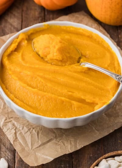 A bowl of smooth pumpkin puree with a spoon on top, placed on parchment paper and a wooden surface. Small pumpkins and seeds are visible in the background.