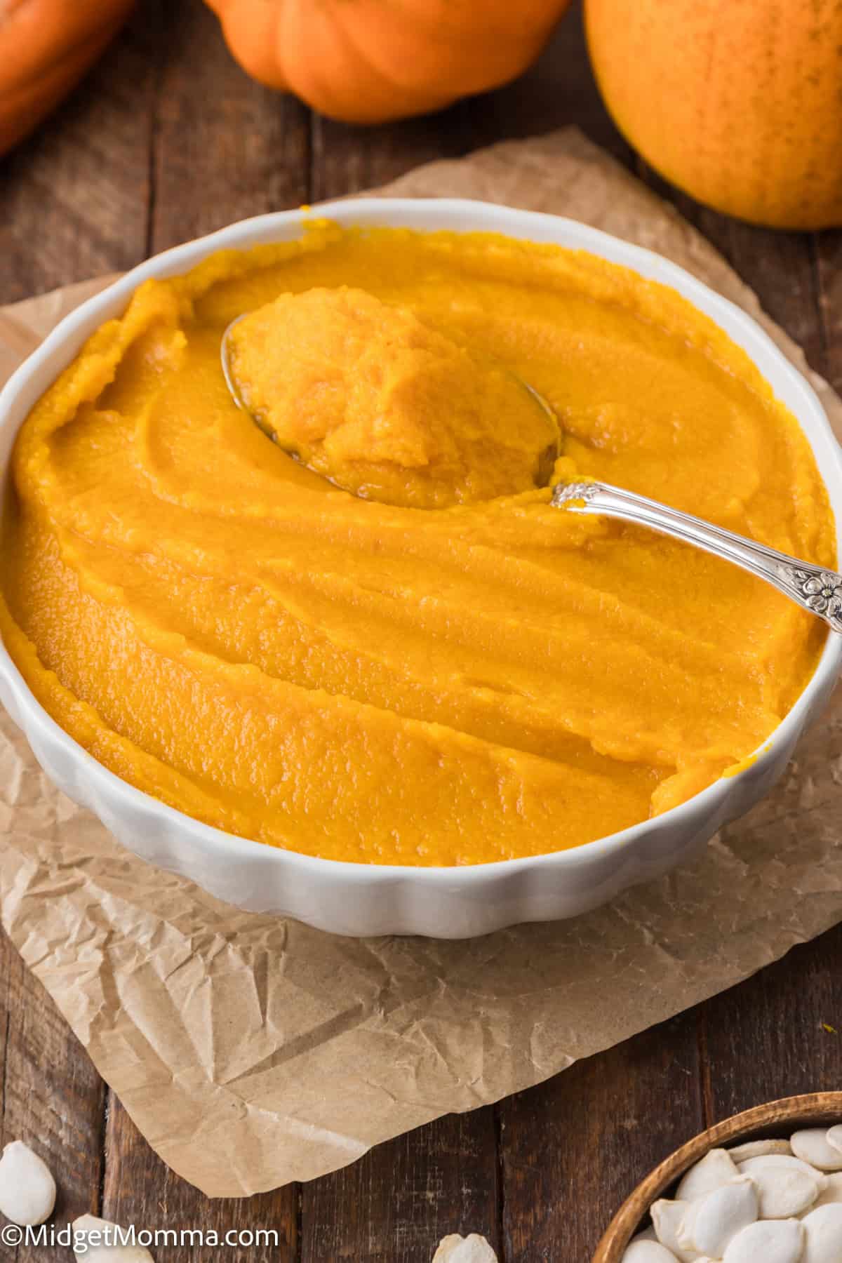 A bowl of smooth pumpkin puree with a spoon on top, placed on parchment paper and a wooden surface. Small pumpkins and seeds are visible in the background.