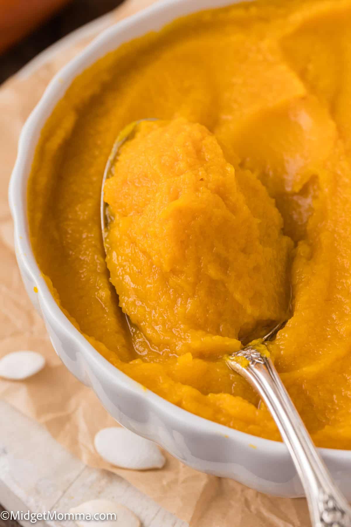 A jar of pumpkin puree with a spoon on a wooden surface, surrounded by pumpkin seeds and a pumpkin slice.