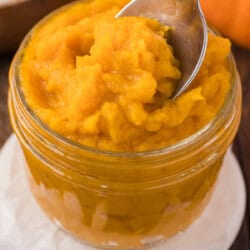 Jar of smooth, bright orange pumpkin puree with a spoon scooping some out. Background shows pumpkin seeds and part of a pumpkin.