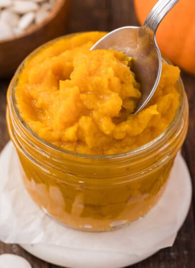 Jar of smooth, bright orange pumpkin puree with a spoon scooping some out. Background shows pumpkin seeds and part of a pumpkin.