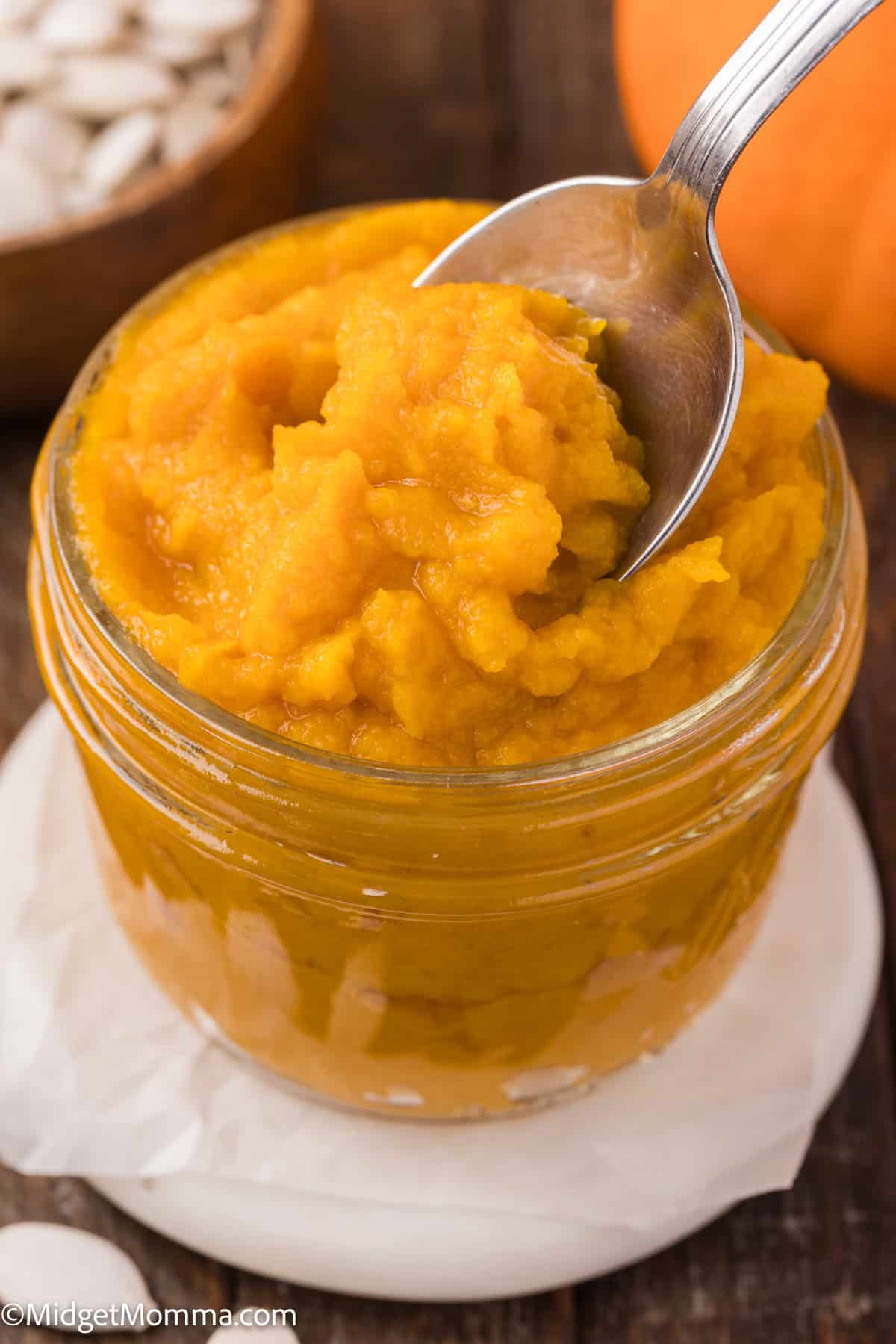 Jar of smooth, bright orange pumpkin puree with a spoon scooping some out. Background shows pumpkin seeds and part of a pumpkin.