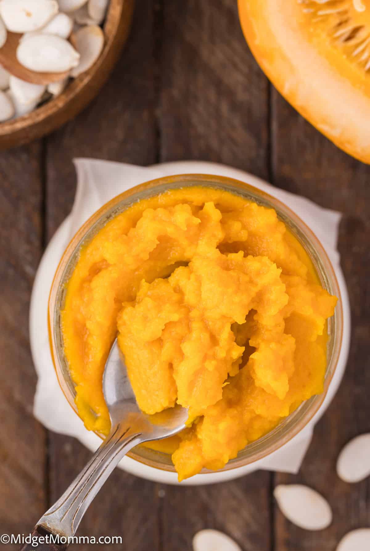 A jar of pumpkin puree with a spoon on a wooden surface, surrounded by pumpkin seeds and a pumpkin slice.