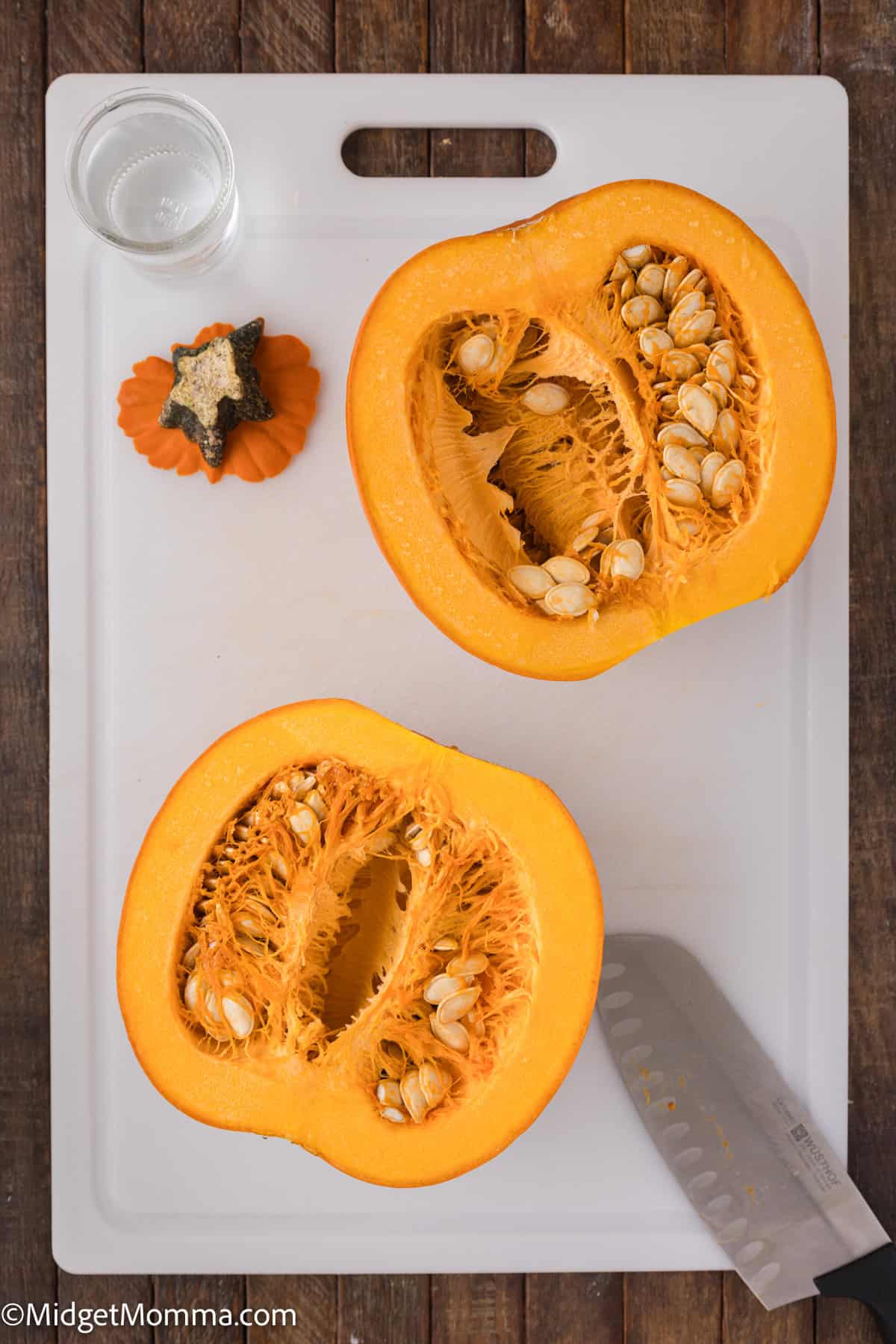 A halved pumpkin on a cutting board with a knife, small decorative pumpkin, and a small glass of clear liquid.