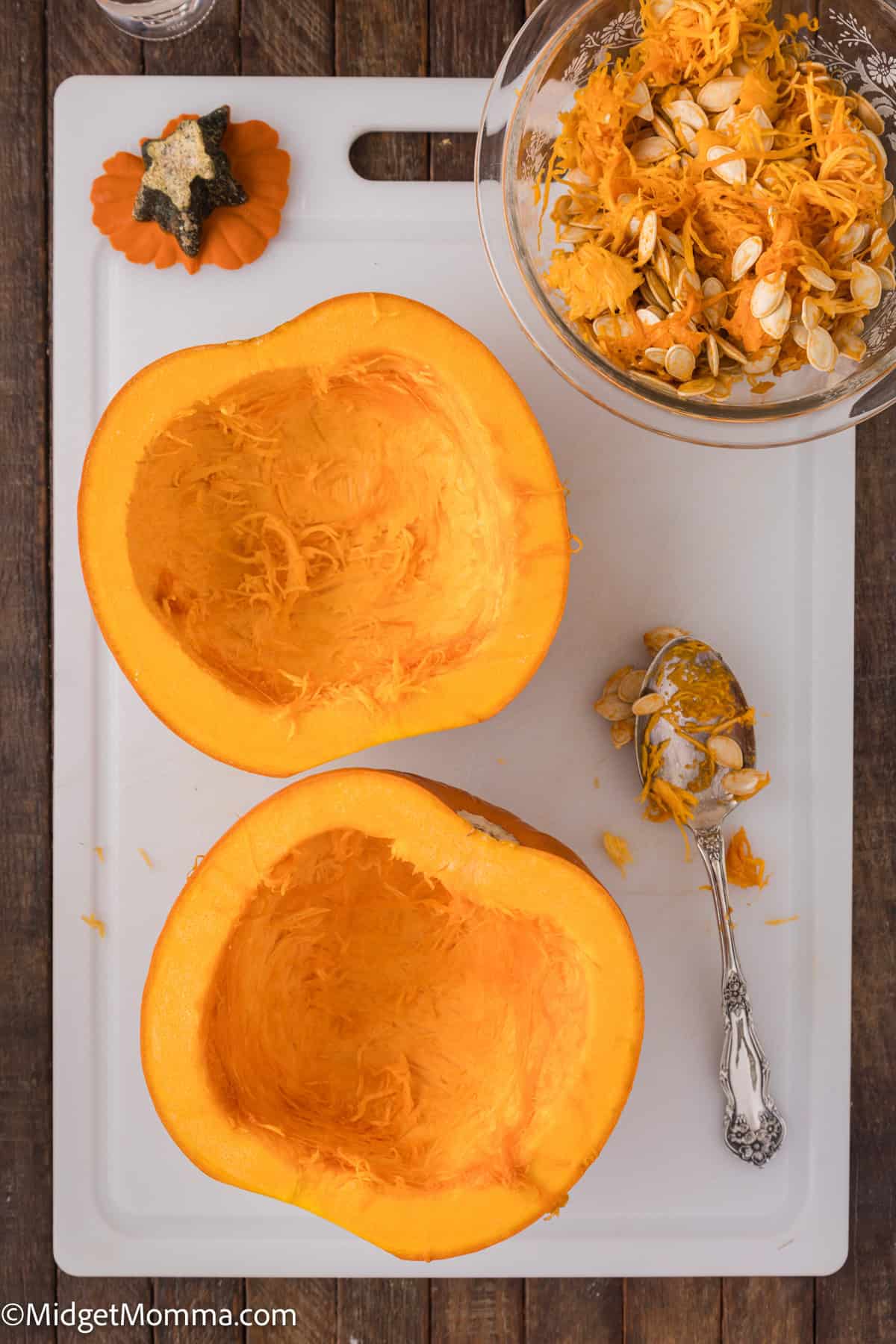 Halved pumpkin on a cutting board with seeds and pulp scooped out into a bowl, next to a spoon and a small decorative pumpkin.