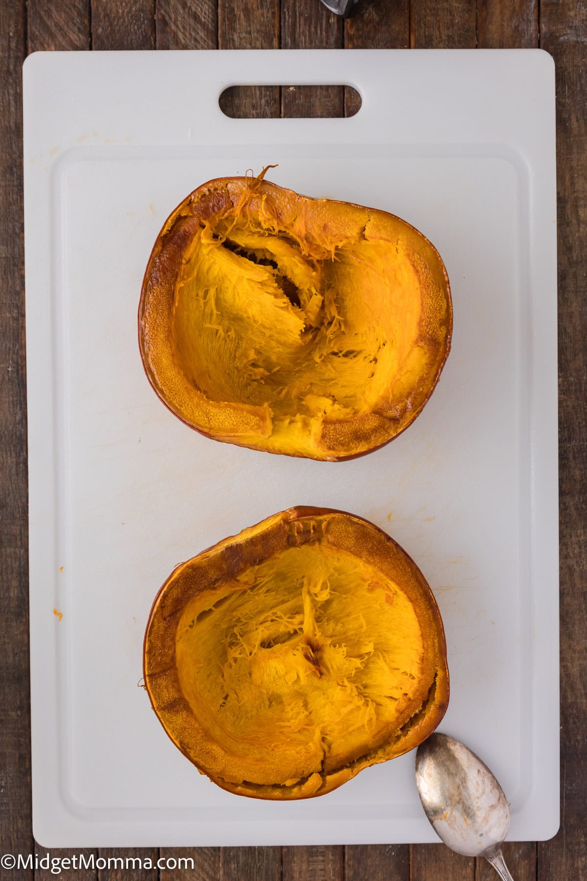 Two halved roasted pumpkins on a white cutting board with a spoon on the side.