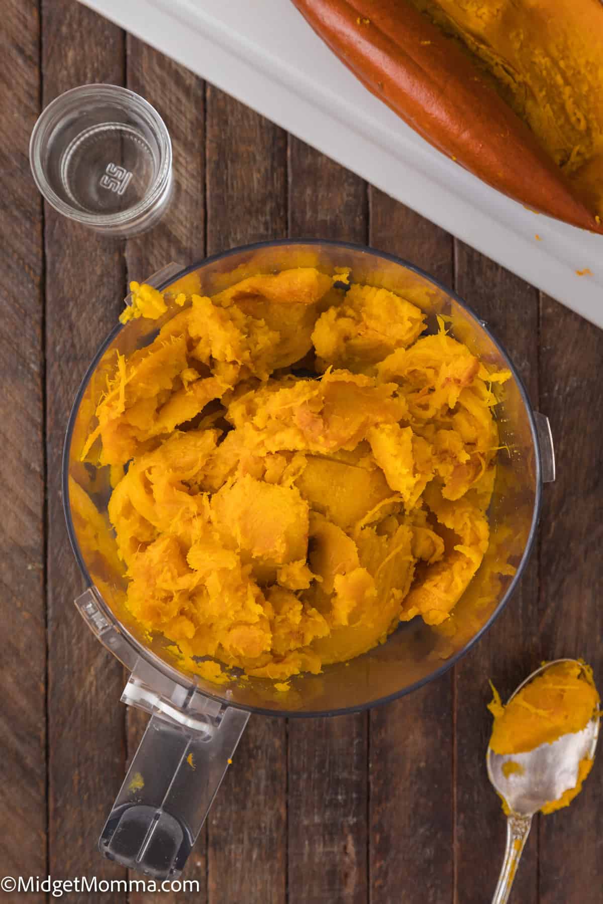 Mashed pumpkin in a food processor bowl on a wooden surface. Nearby are a spoon, a glass cup, and a partially used pumpkin on a cutting board.