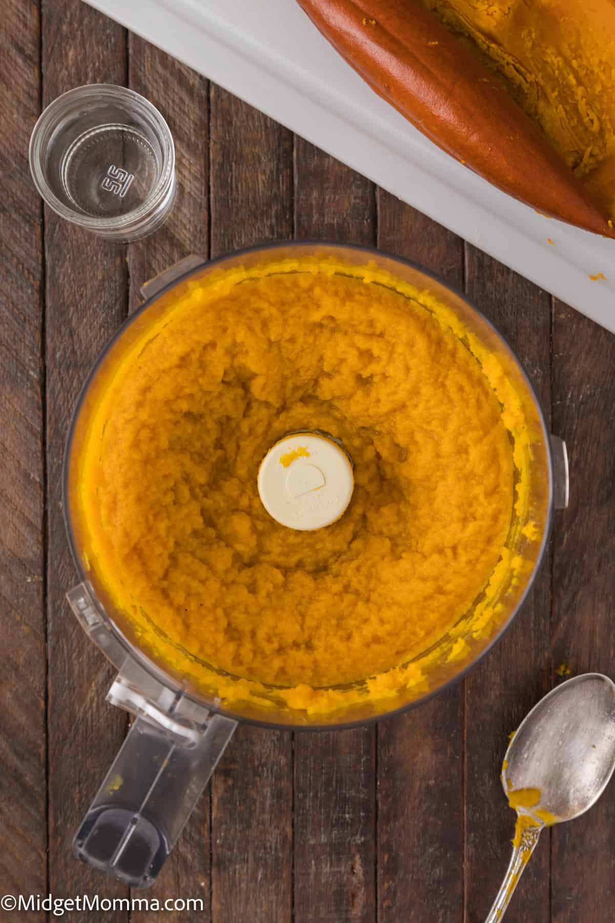 A food processor containing blended pumpkin is on a wooden table. A spoon and a small glass sit nearby.