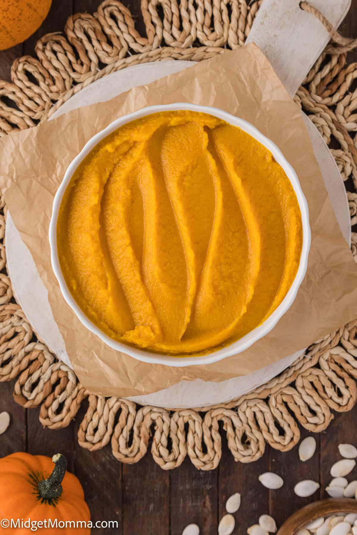 Bowl of smooth orange pumpkin puree on a wooden board with a woven mat underneath, surrounded by pumpkin seeds.