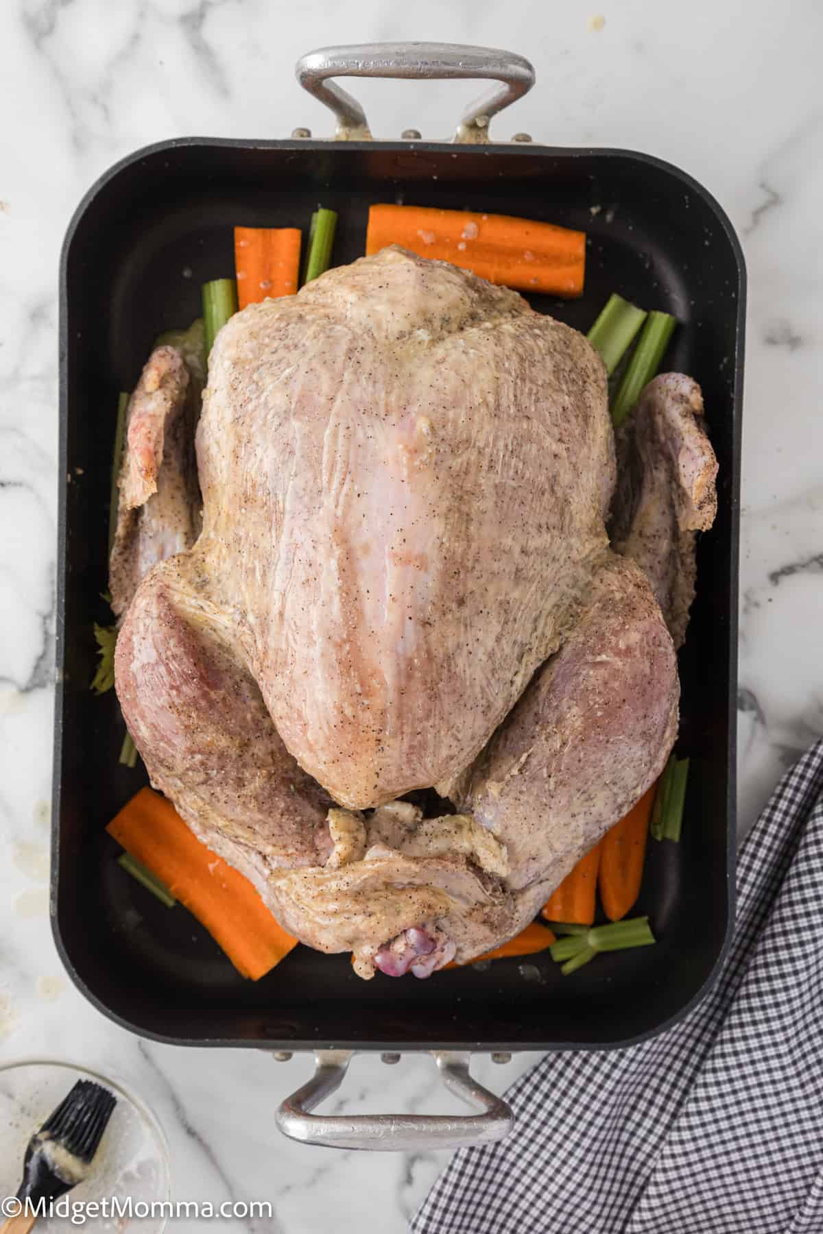 Whole raw turkey on a bed of carrots and celery in a roasting pan, placed on a marble surface.