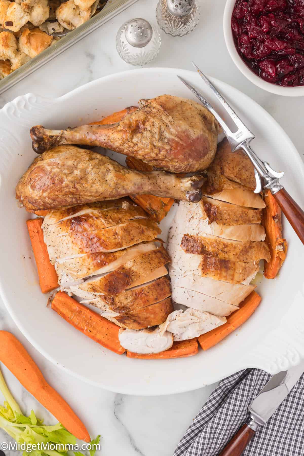 Sliced turkey breast and drumsticks on a white plate with carrots, next to a dish of cranberry sauce and a casserole dish.