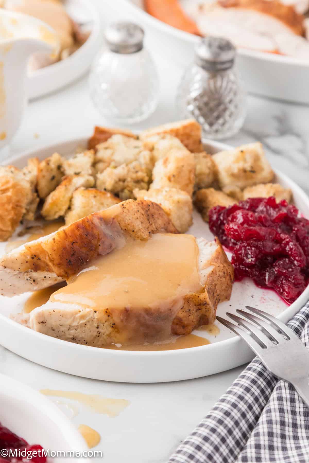 Plate with sliced turkey covered in gravy, stuffing, and cranberry sauce on a table with salt and pepper shakers and a checkered napkin.