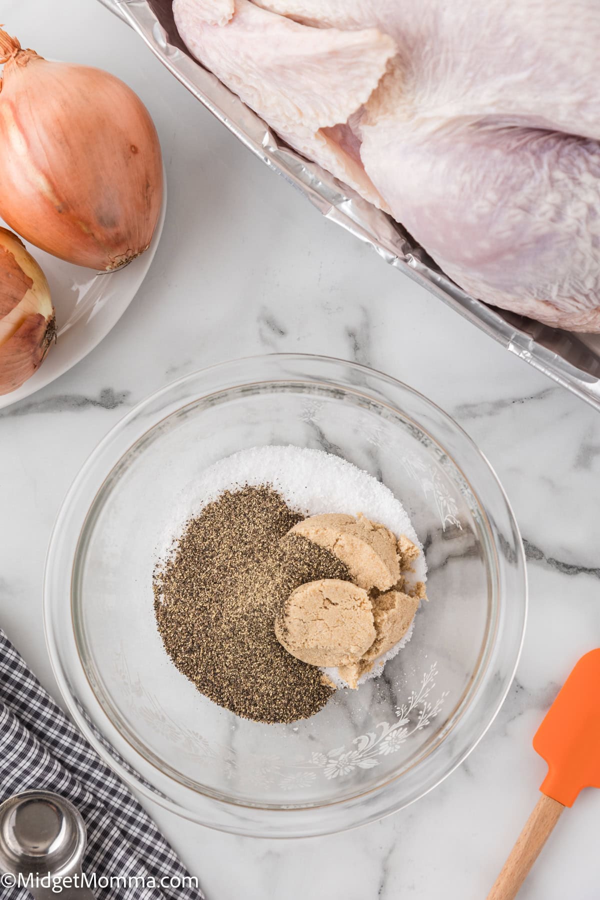 A bowl with brown sugar, salt, and pepper mix on a marble countertop for the Dry Brine Turkey Recipe. Nearby are onions, a turkey, a gray towel, and an orange spatula.