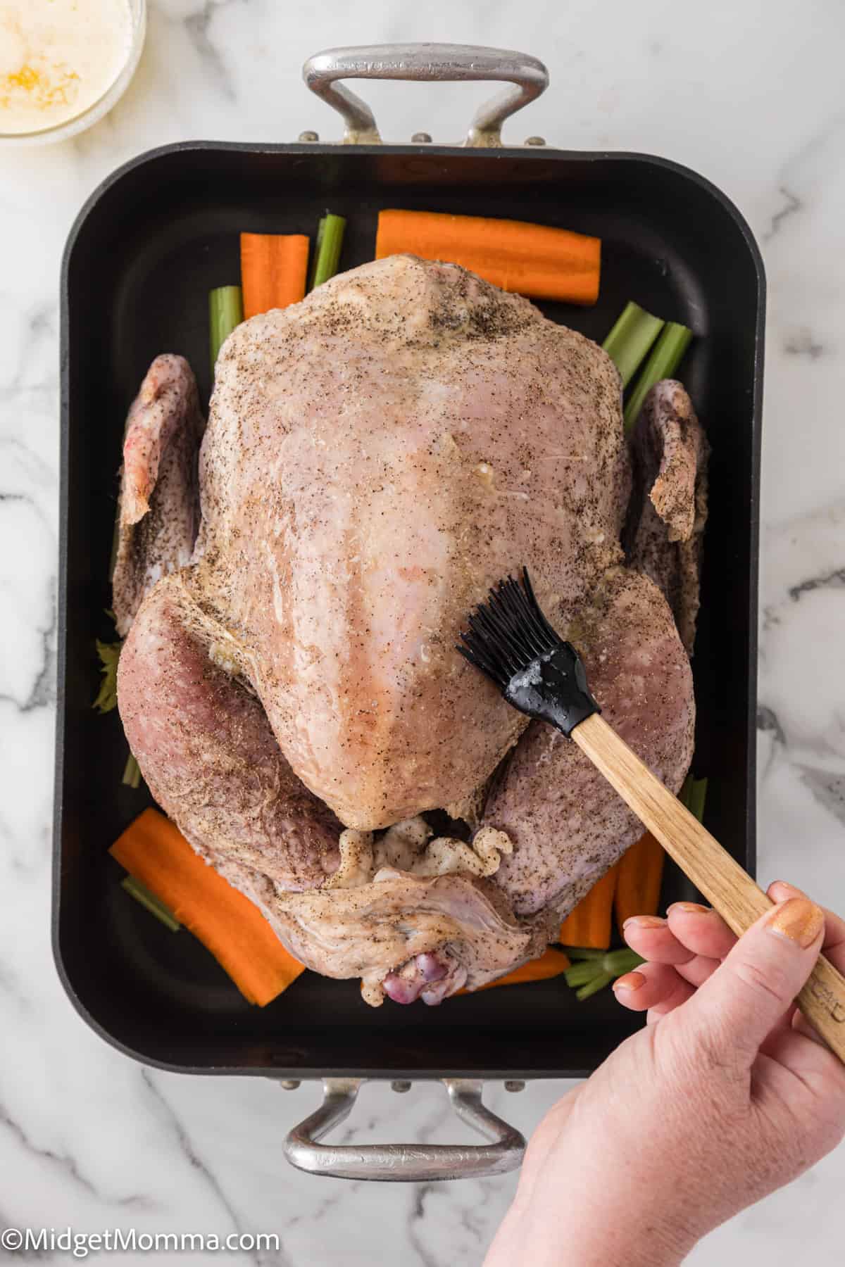 A raw turkey in a roasting pan is being brushed with seasoning. Carrot and celery pieces are placed around the turkey on a marble countertop.