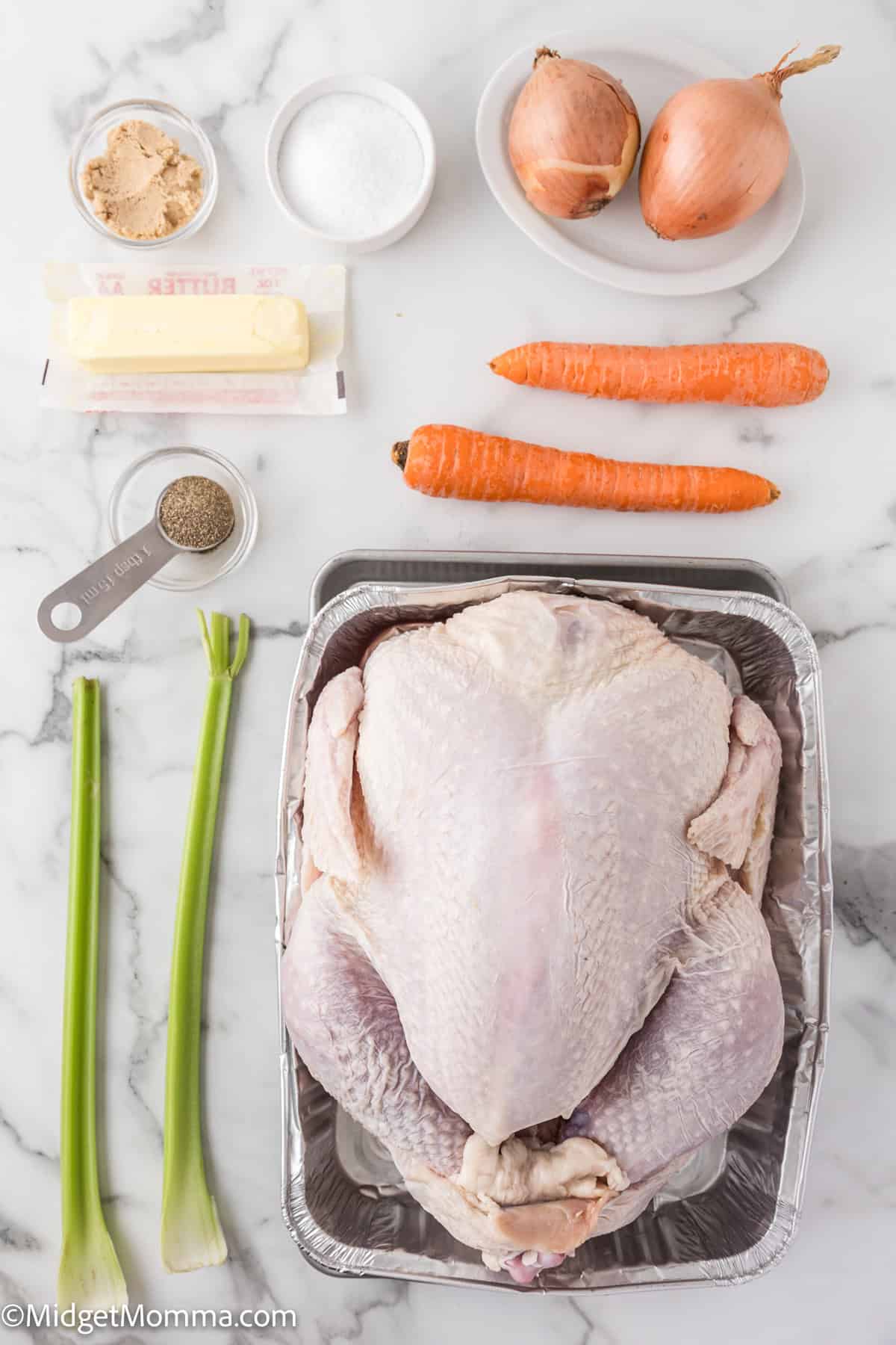 Raw whole chicken in a pan with ingredients including carrots, onions, celery, butter, brown sugar, salt, and pepper arranged on a marble surface.