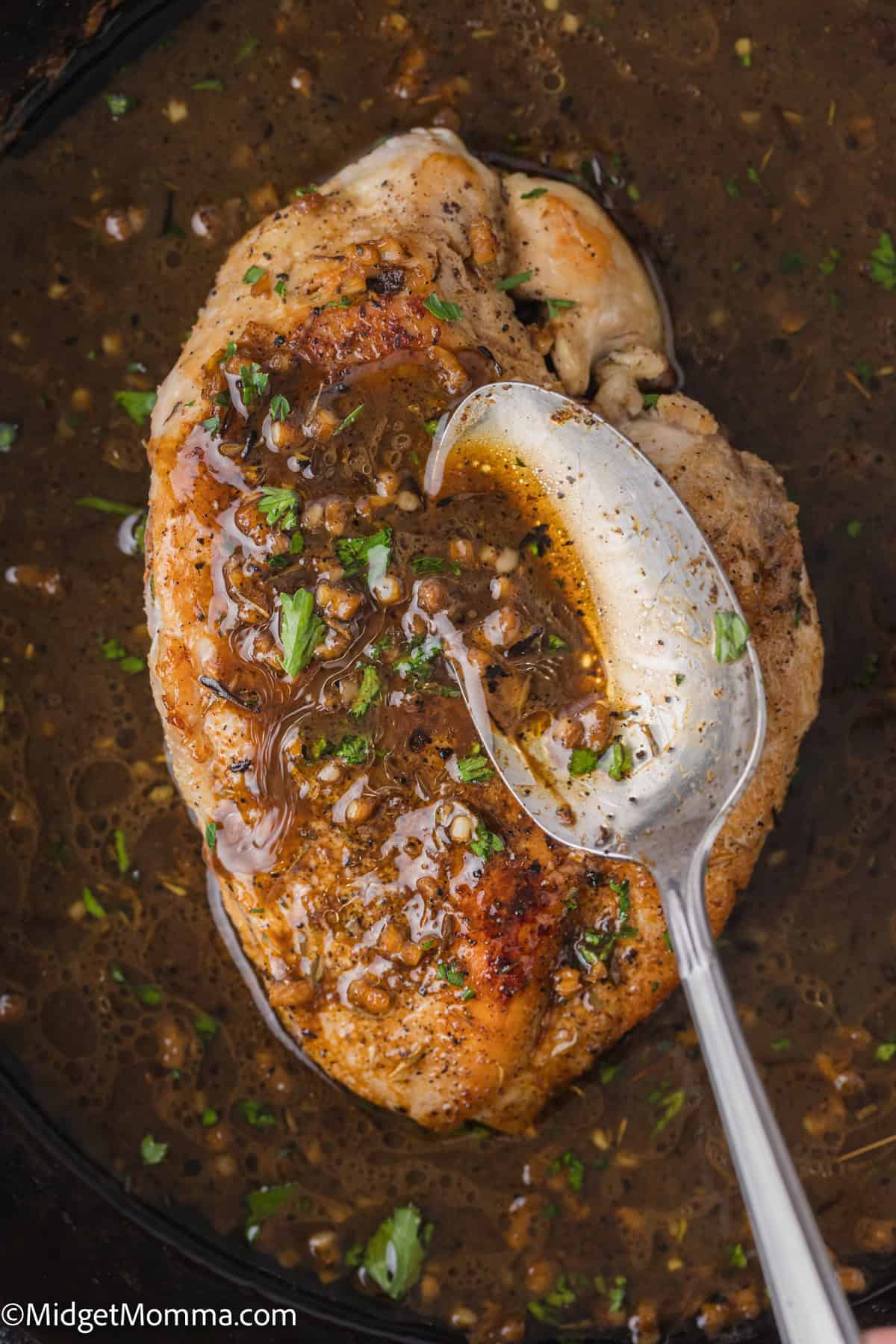 A cooked chicken breast covered in a brown sauce with seasoning and herbs, with a spoon placed on top of it, in a dark pan.