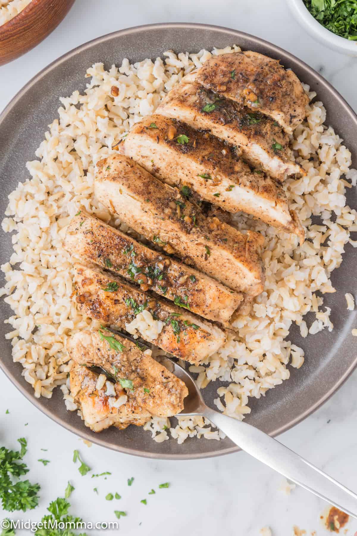 A plate with sliced seasoned chicken breast served on a bed of rice. A fork is placed beside the chicken. Fresh herbs are sprinkled on top.