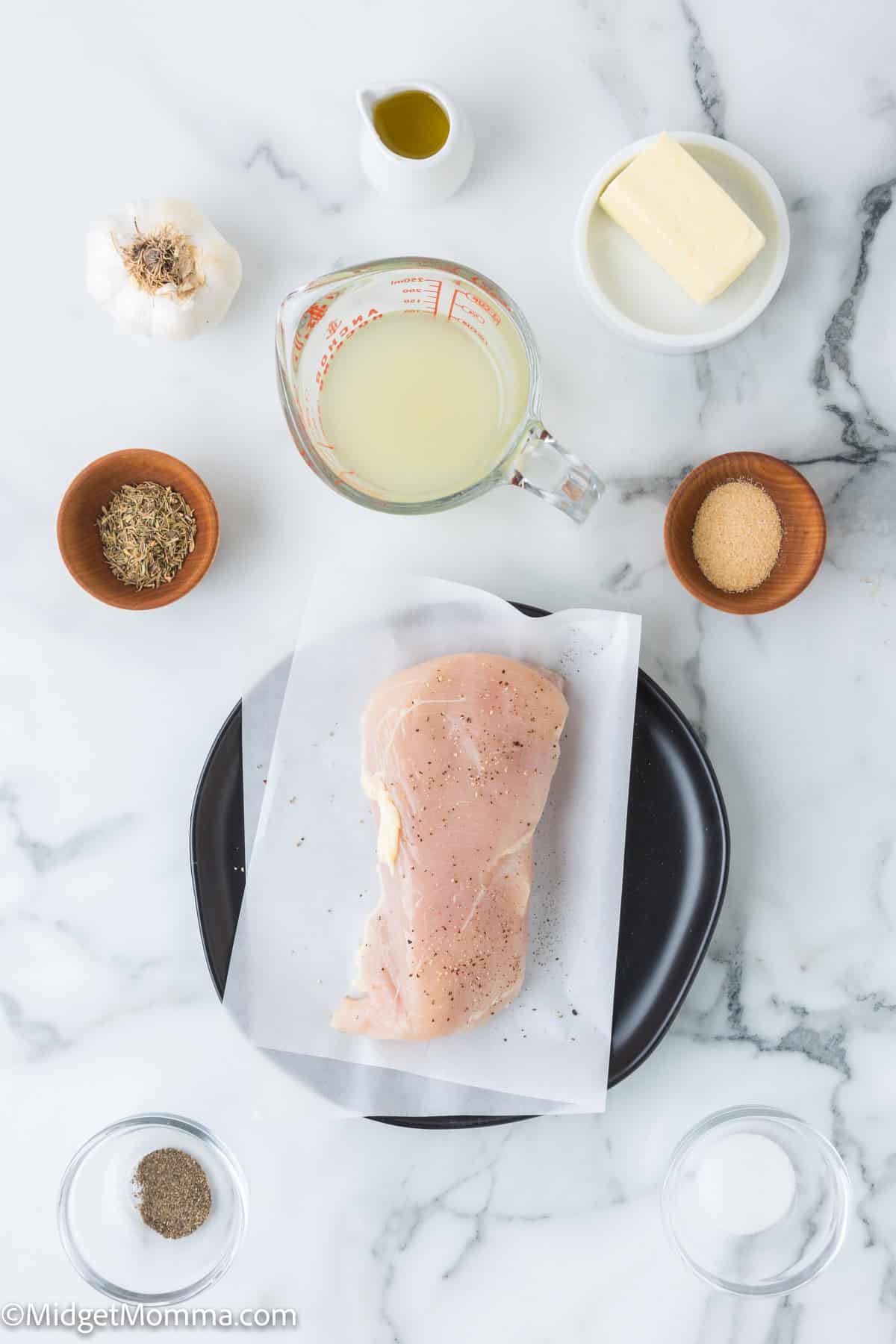 Garlic Butter Chicken Ingredients - A raw seasoned chicken breast on a plate surrounded by ingredients including garlic, olive oil, butter, lemon juice, spices, and seasonings on a marble surface.