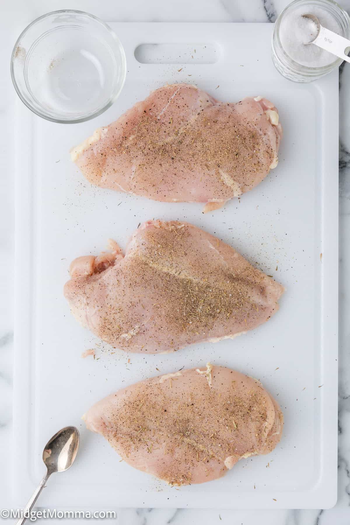 Three raw chicken breasts seasoned with spices are placed on a white cutting board. A glass bowl, a measuring spoon, and a salt shaker are also visible on the board.
