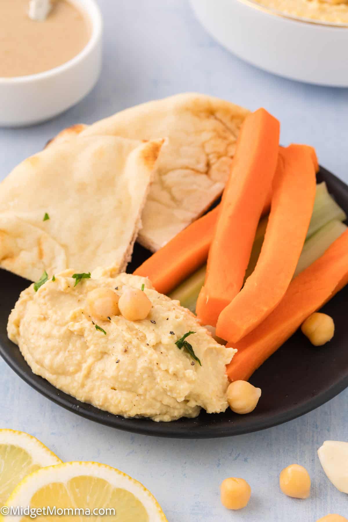 A bowl of creamy hummus topped with whole chickpeas, a sprig of parsley, and a drizzle of olive oil. A wooden-handled spoon rests on the right side of the bowl.