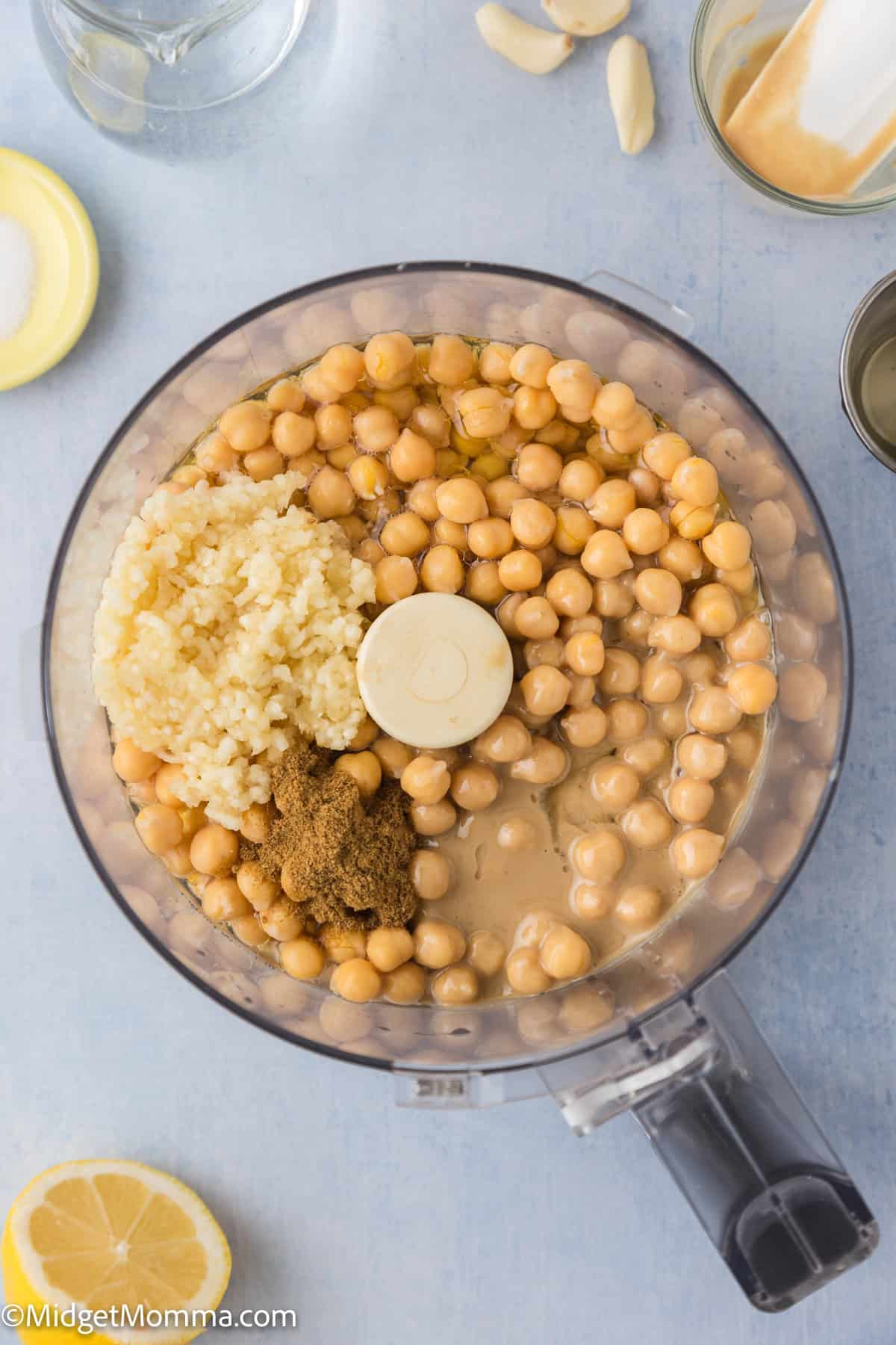 A food processor filled with chickpeas, minced garlic, ground spices, lemon juice, and liquid ingredients, viewed from above.