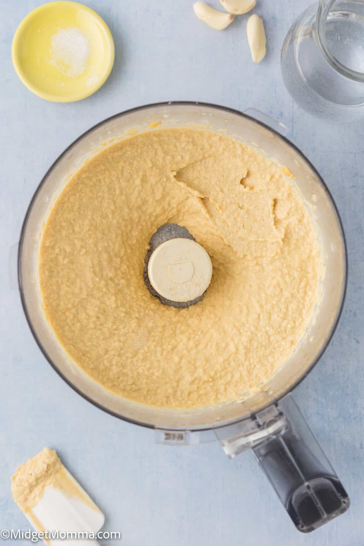 Image of hummus being prepared in a food processor. Nearby are a small bowl of salt, a few garlic cloves, and a glass of water.