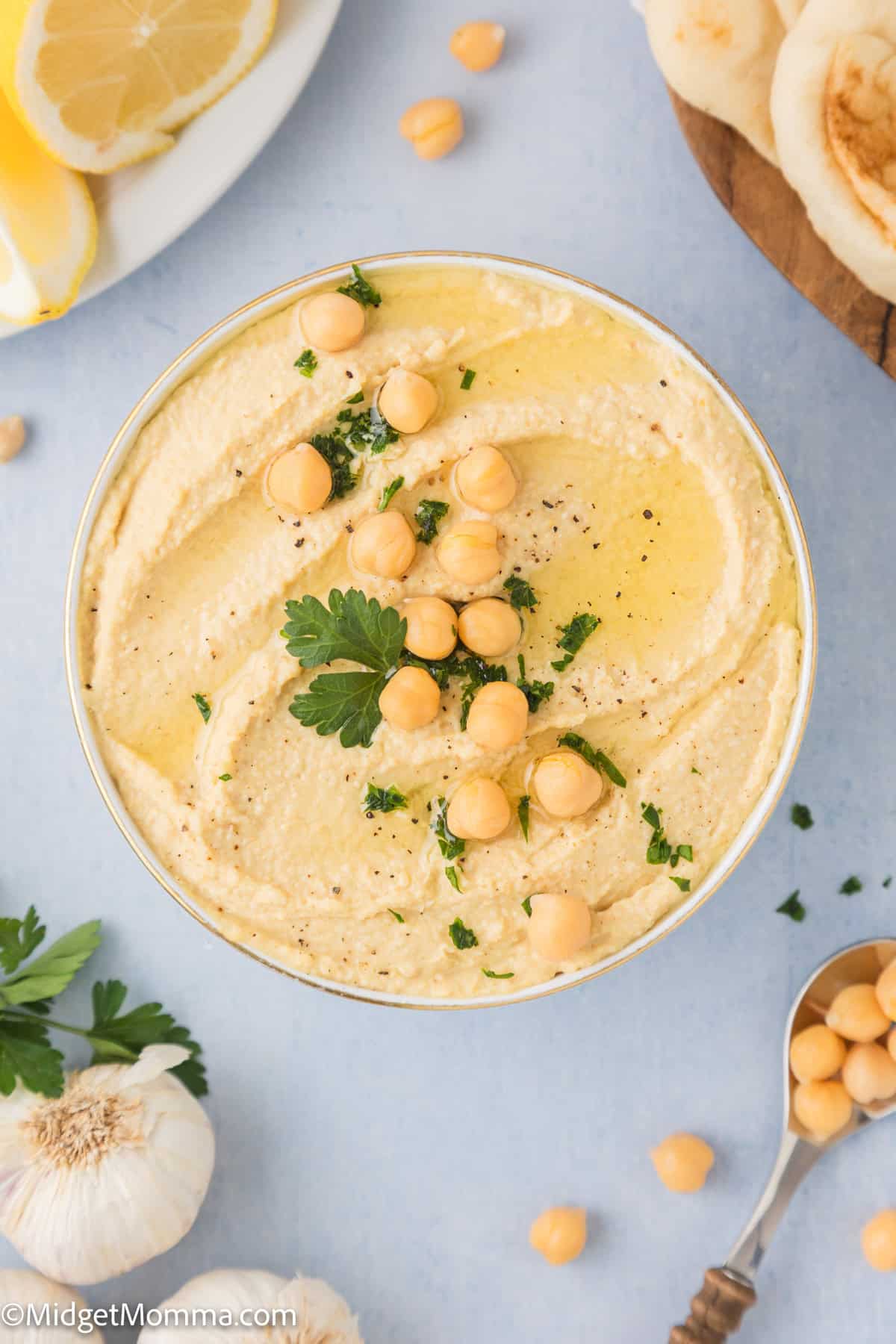 A bowl of hummus garnished with chickpeas and parsley, surrounded by sliced lemons, pita bread, a garlic bulb, and a spoon with chickpeas.