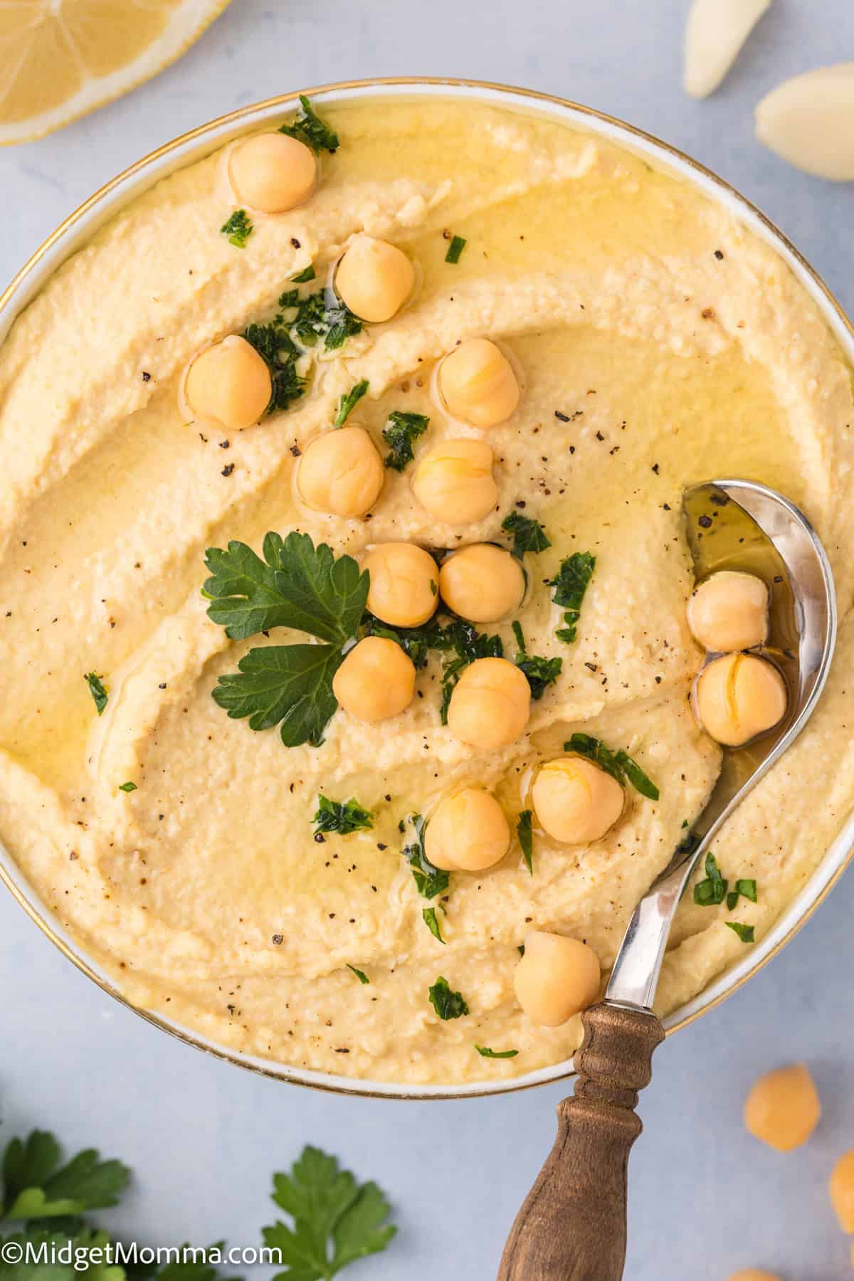 A bowl of creamy hummus topped with whole chickpeas, a sprig of parsley, and a drizzle of olive oil. A wooden-handled spoon rests on the right side of the bowl.