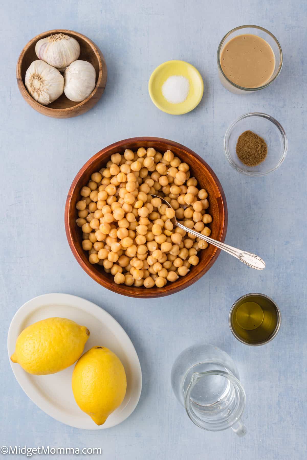 Ingredients for making hummus include a bowl of chickpeas, garlic, salt, cumin, tahini, olive oil, lemons, and water.