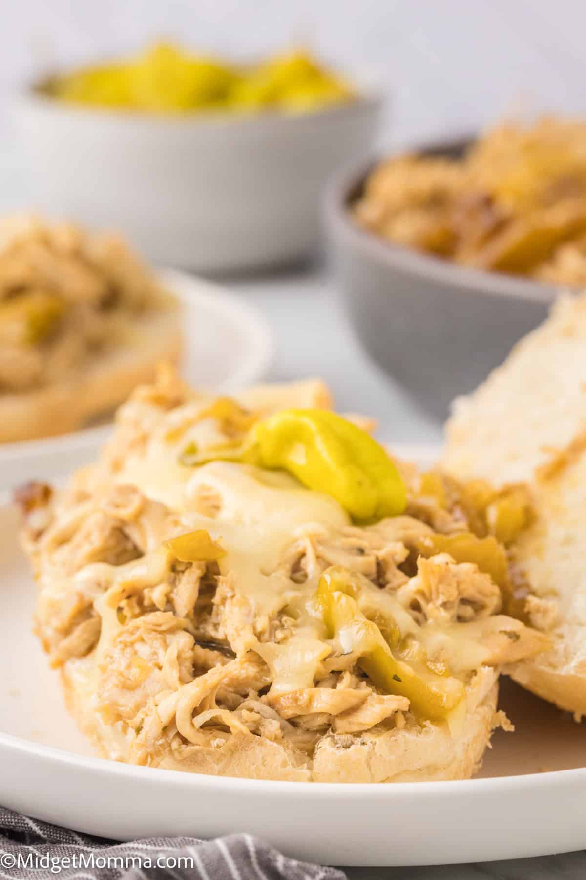 close up photo of a A mississippi chicken sandwich with shredded chicken and melted cheese topped with a pepperoncini on a white plate, accompanied by a bowl of more chicken and peppers on the side.
