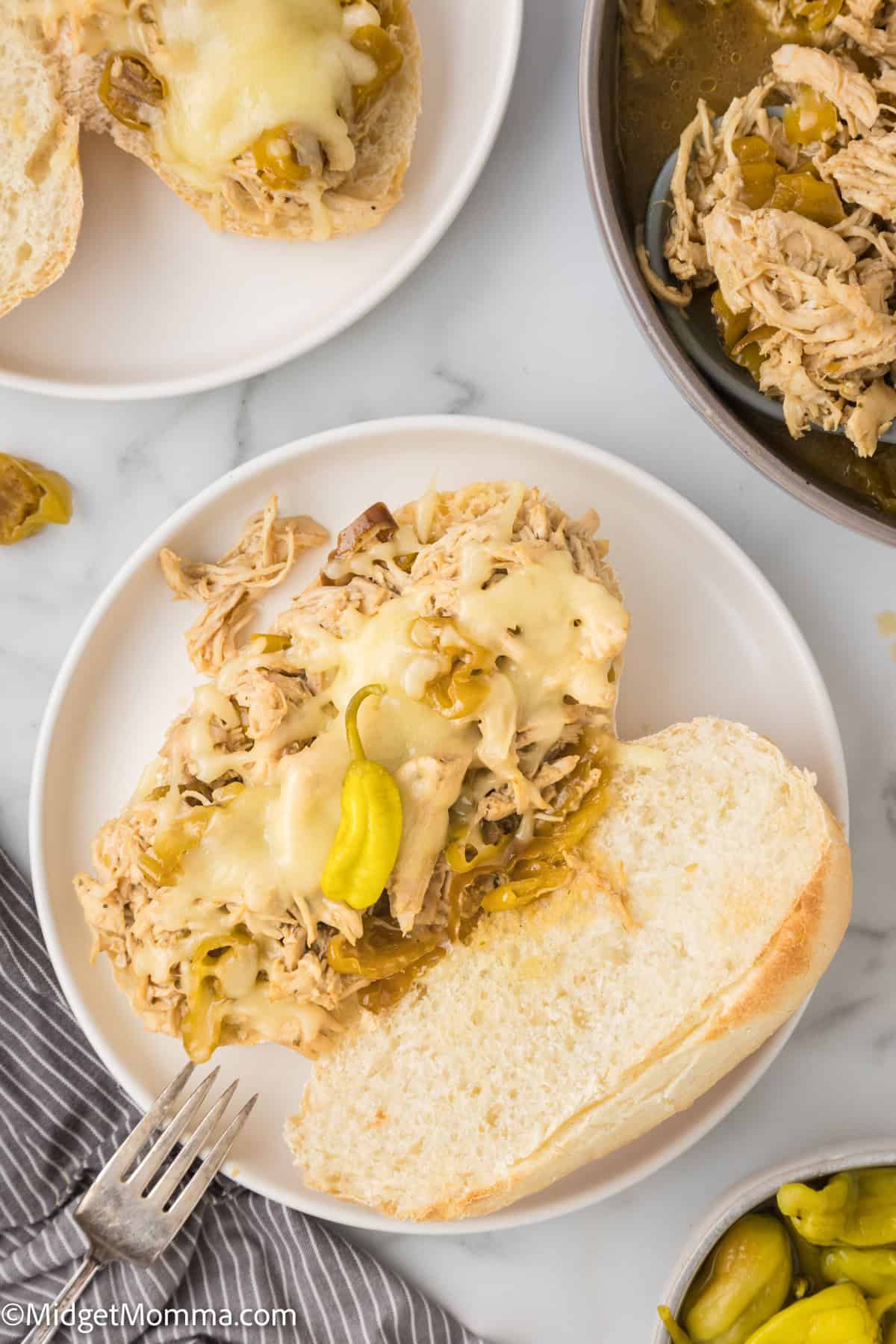A mississippi chicken sandwich with shredded chicken and melted cheese topped with a pepperoncini on a white plate, accompanied by a bowl of more chicken and peppers on the side.