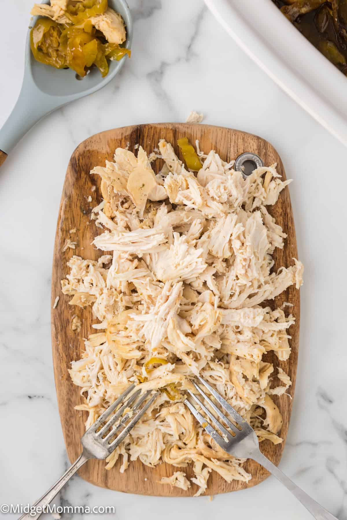 Shredded chicken on a wooden cutting board with forks, accompanied by a side bowl of pickles.