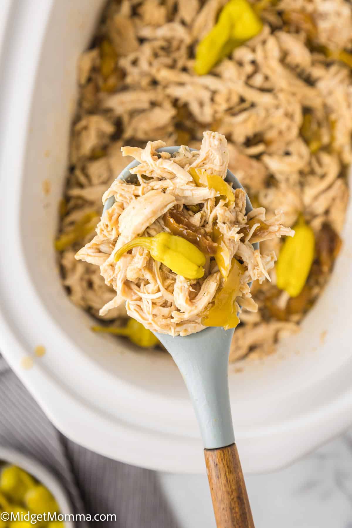 A spoon lifting shredded chicken with peppers from a slow cooker.