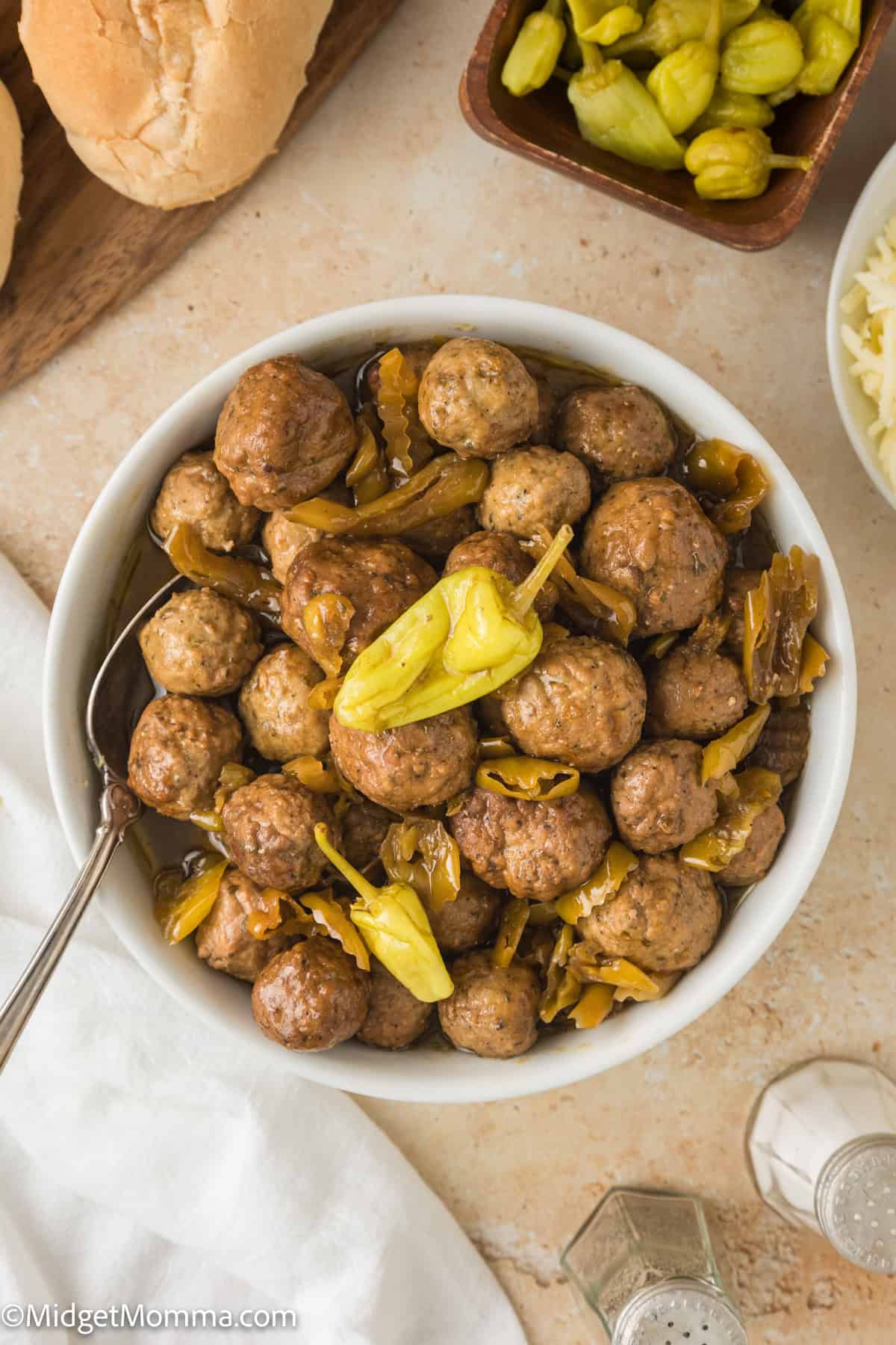 A bowl filled with meatballs and pepperoncini peppers, serving utensils, and a roll of bread beside it.