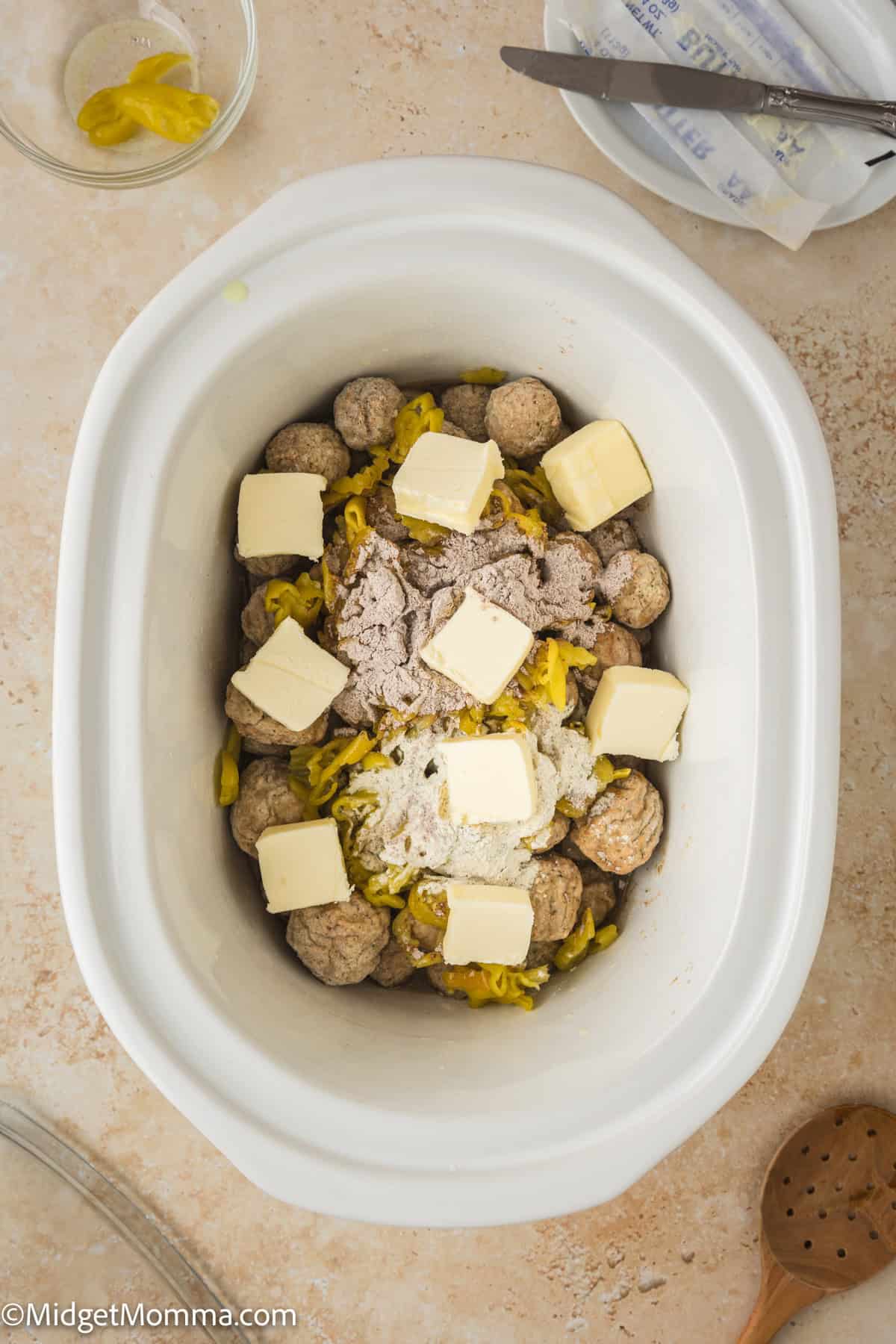A slow cooker filled with meatballs, sliced yellow peppers, seasonings, and cubes of butter, ready to be cooked. A wooden spoon and a stick of butter are visible nearby.
