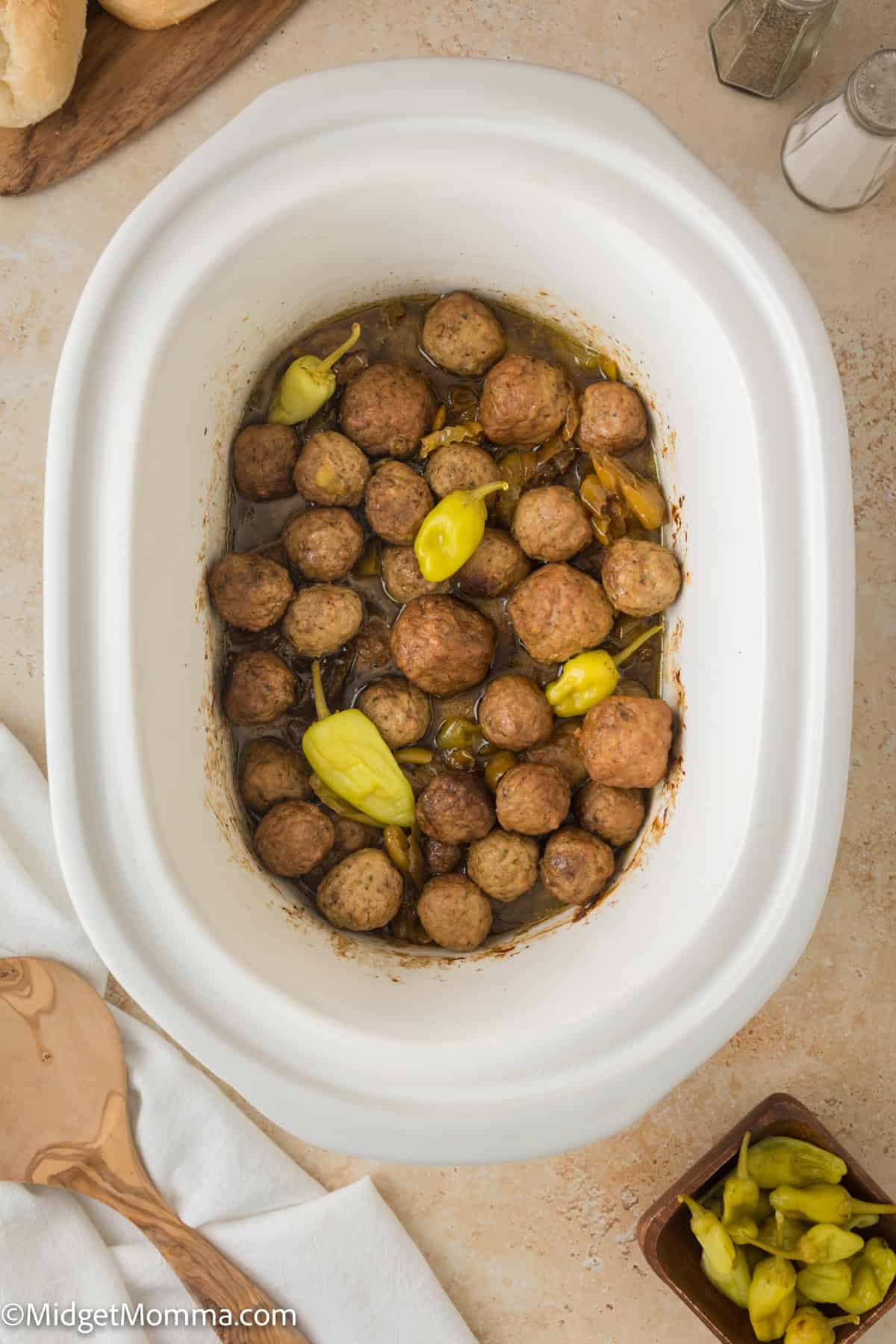 A white slow cooker filled with cooked meatballs and several whole pepperoncini peppers. A wooden spoon and a small bowl of additional pepperoncini are nearby.