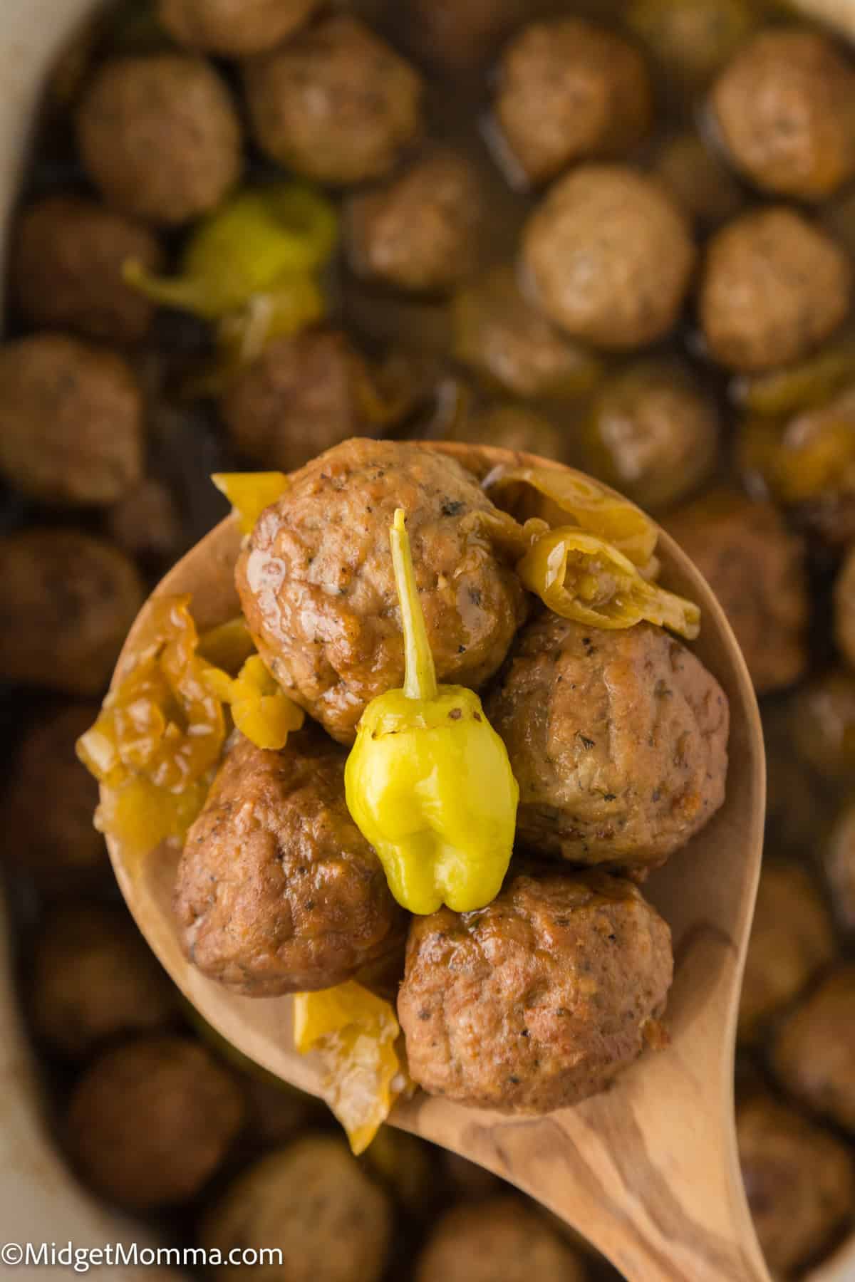 A wooden spoon holds several meatballs and a pepperoncini pepper above a pot full of more meatballs.