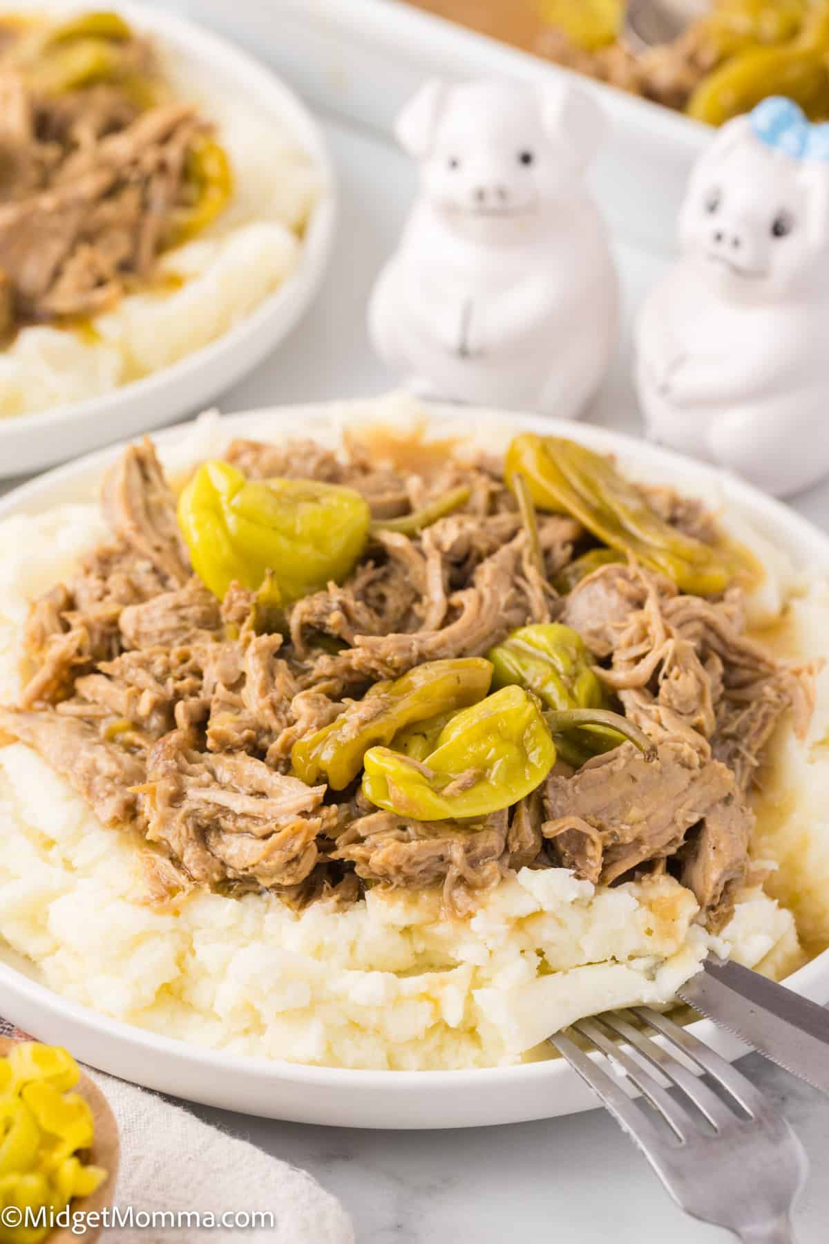 A plate of shredded pork roast topped with peppers served on a bed of mashed potatoes. Two pig-shaped salt and pepper shakers are in the background. A fork is resting on the plate.