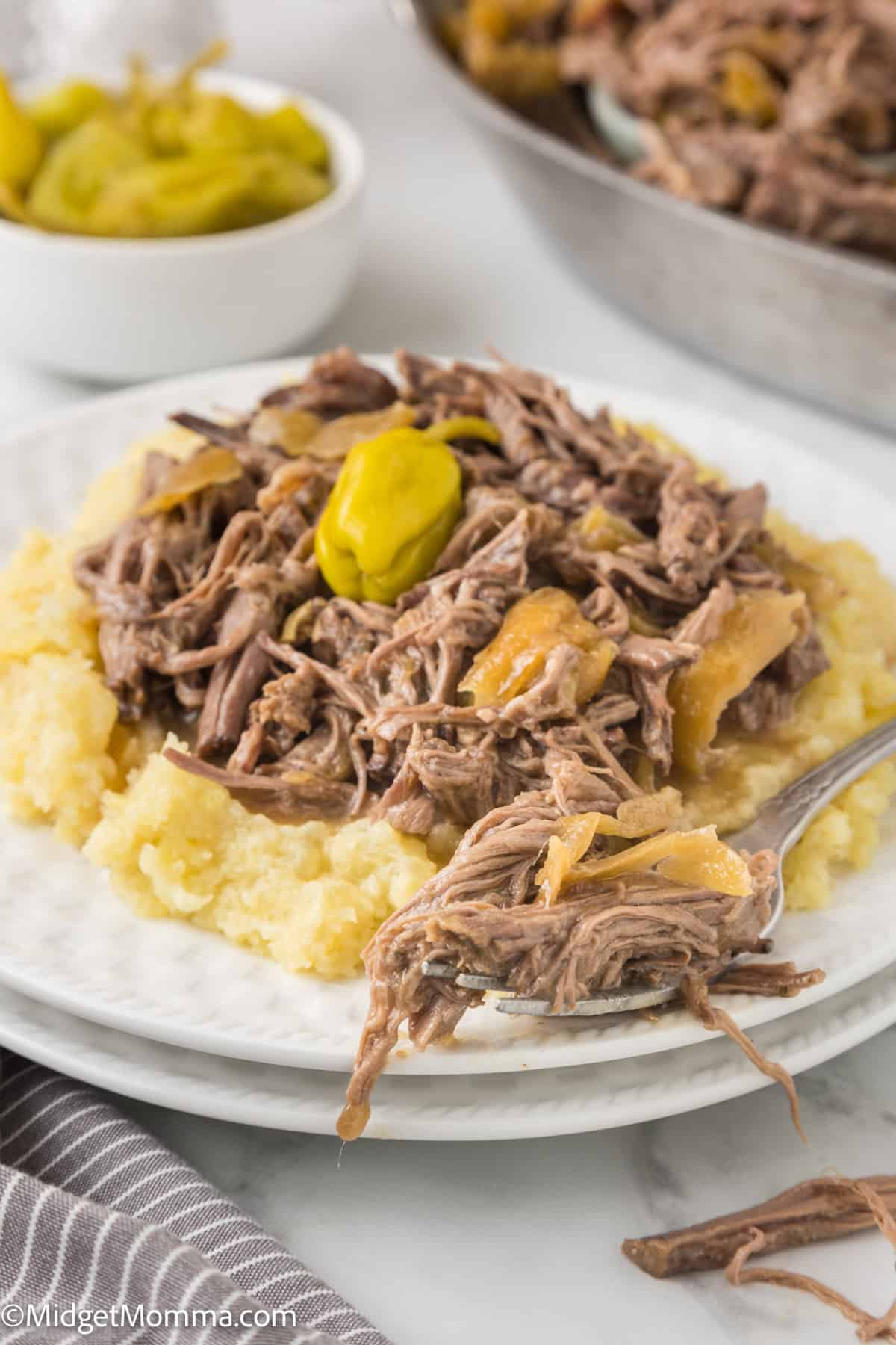 A plate of shredded mississippi pot roast beef served on mashed potatoes with peppers on top. A fork rests beside the dish.