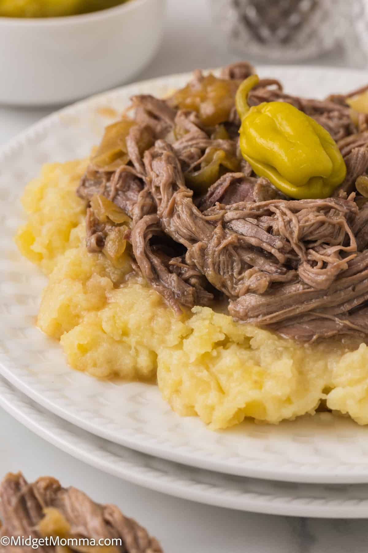 A plate with mashed potatoes topped with shredded beef and a green pepper. A bowl with more peppers is partially visible in the background.