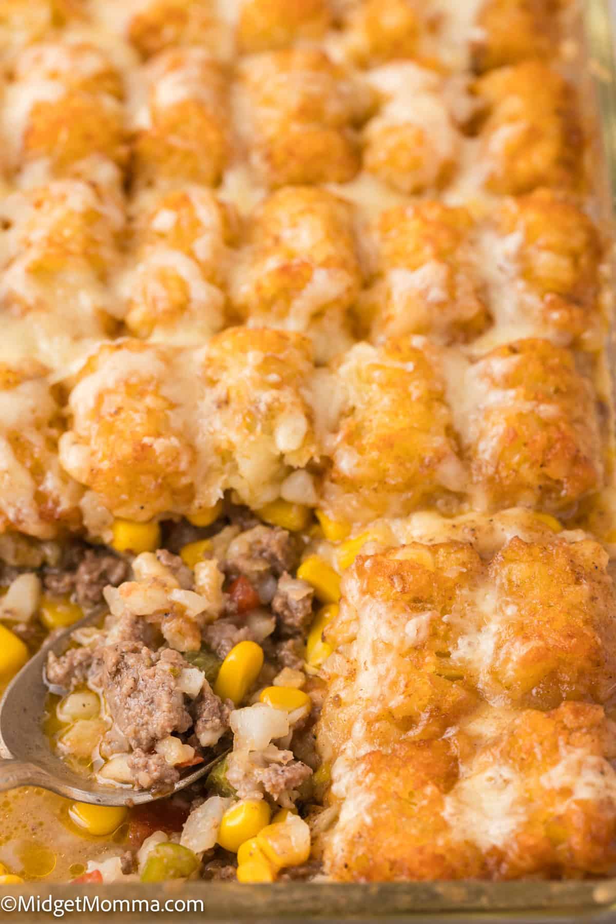 A baked casserole dish with tater tots on top, a scoop revealing ground beef, corn, and other ingredients underneath.