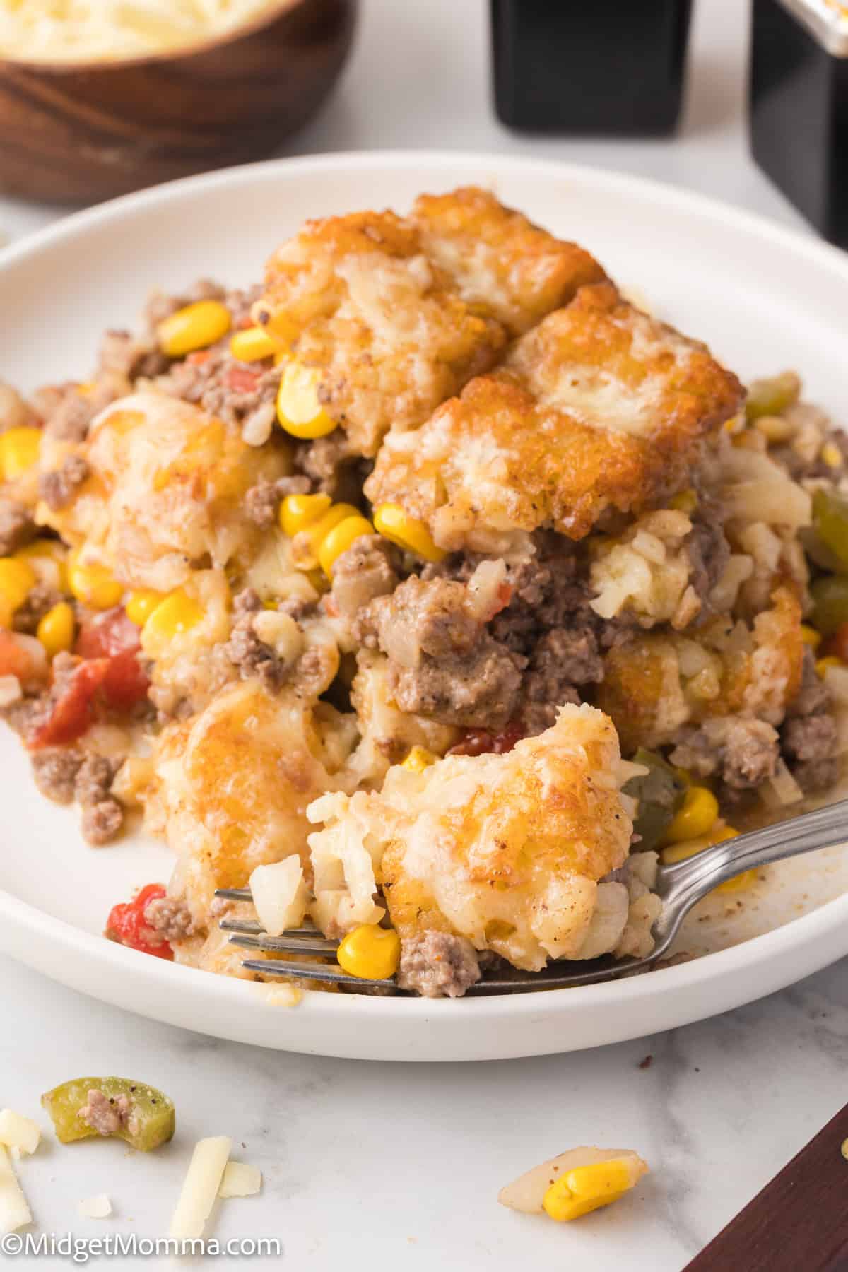 A close-up of a plate of tater tot casserole with ground beef, corn, and other vegetables. A fork is placed on the plate, partially underneath a portion of the casserole.