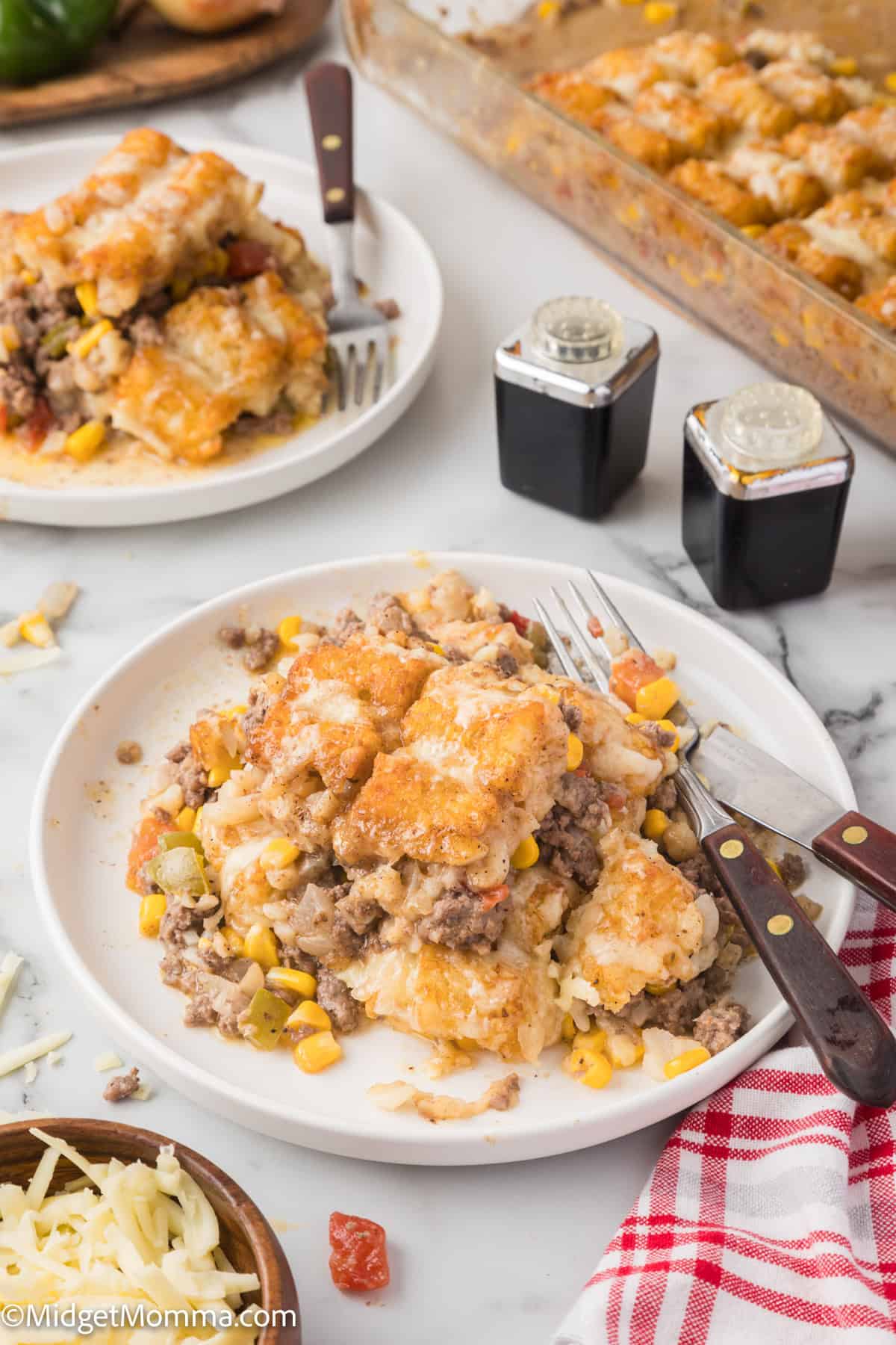 A plate with a serving of a casserole containing ground meat, corn, and a biscuit topping. Two forks are placed on the plate. A pan with more casserole and condiments are in the background.